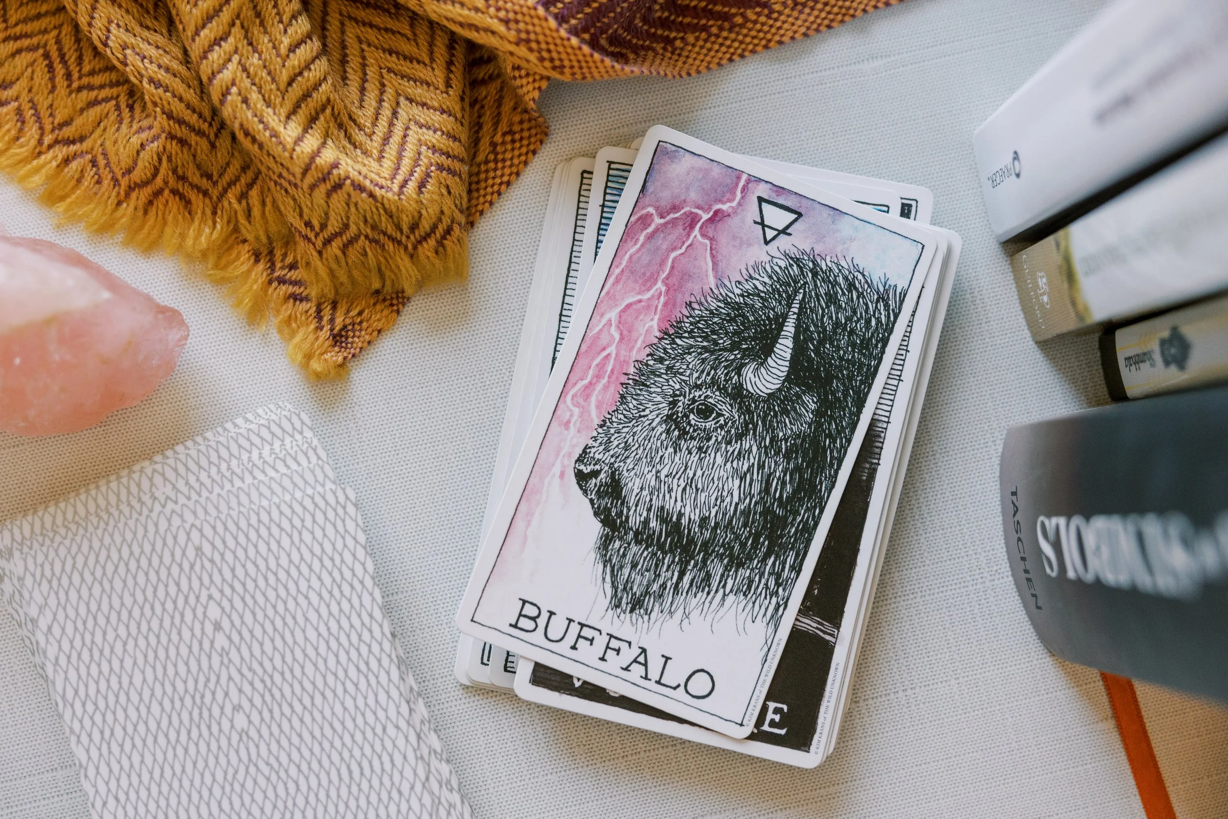 A deck of cards featuring illustrations of animals with one card showing a buffalo, with a background of pink and lightning. The cards are on a white surface alongside a gold-patterned cloth, a salt lamp, and stacked books.