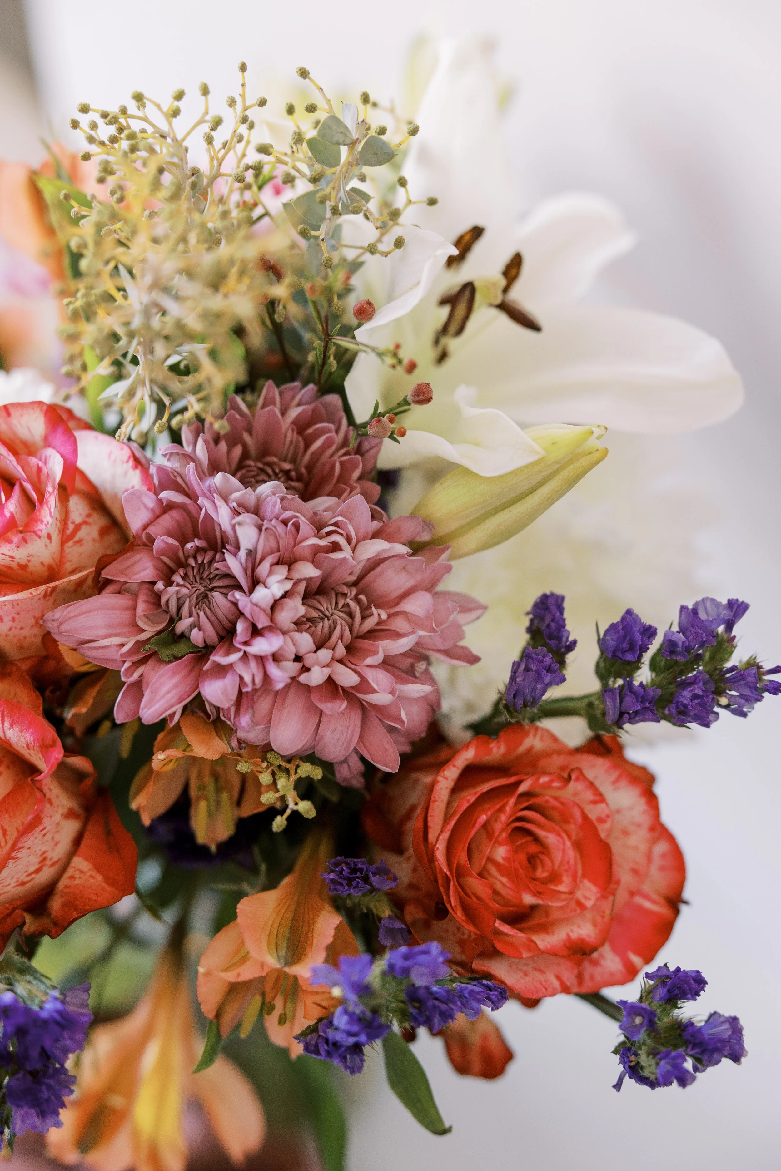 Close-up of a colorful bouquet of flowers including pink dahlias, orange roses, white lilies, purple statice, and various greenery.