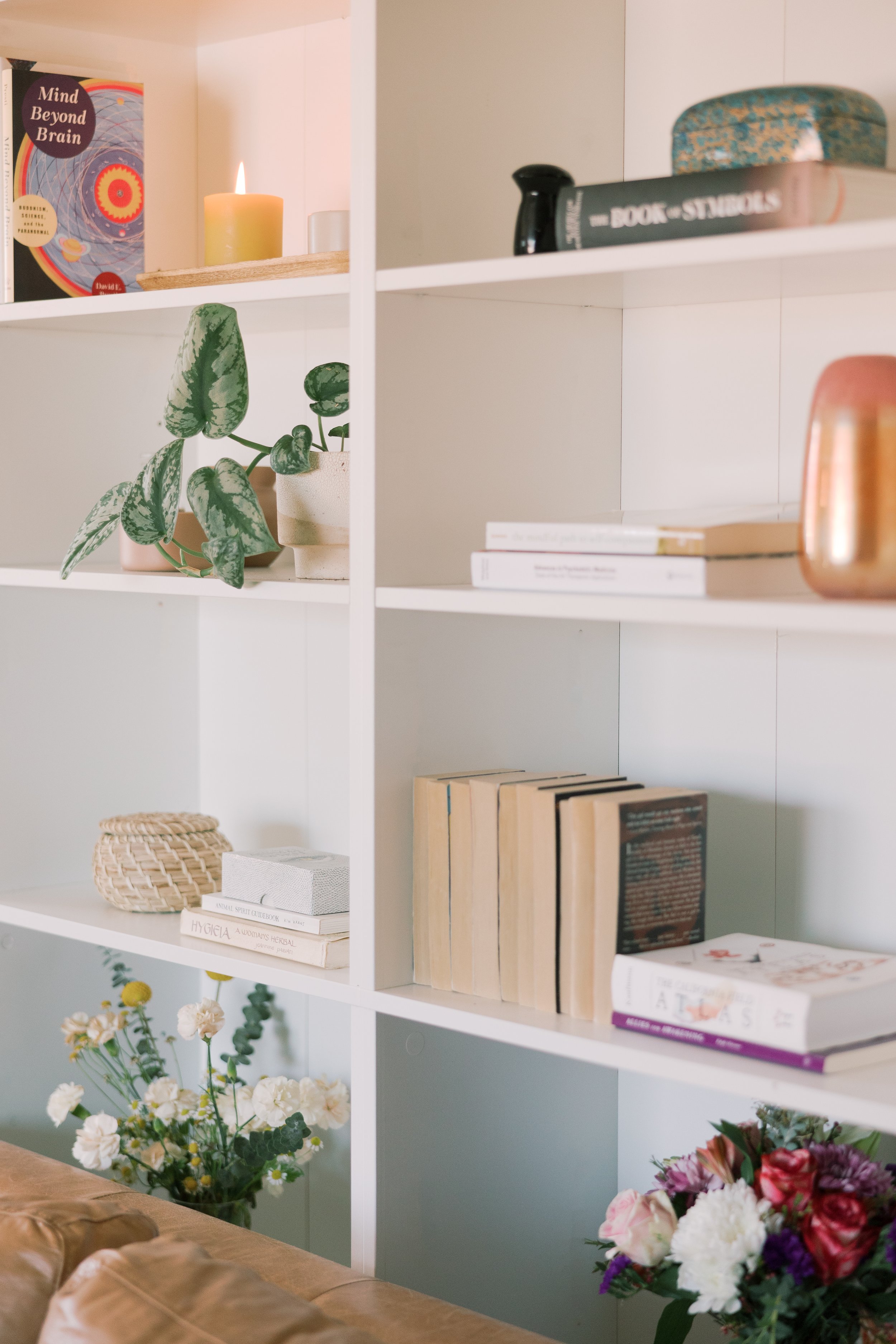 White bookshelf with books, potted plants, candles, and decorative items in a cozy room.