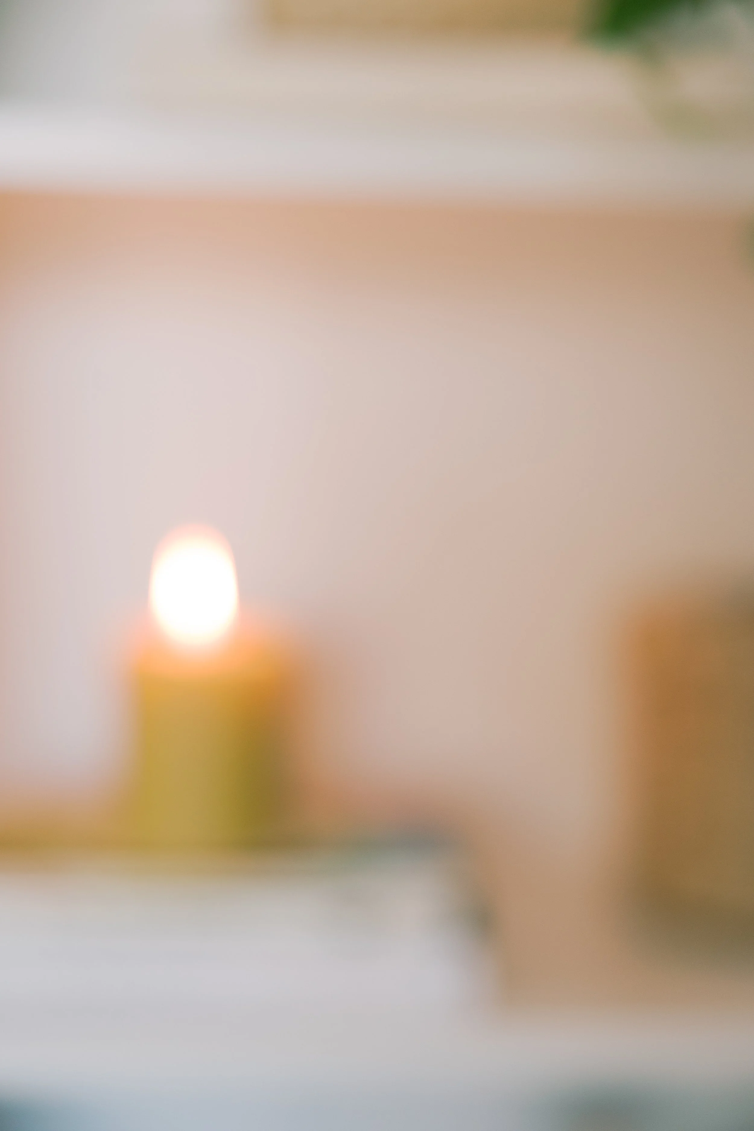 A blurred image of a yellow candle with a lit flame, placed on a white surface, with a beige background.