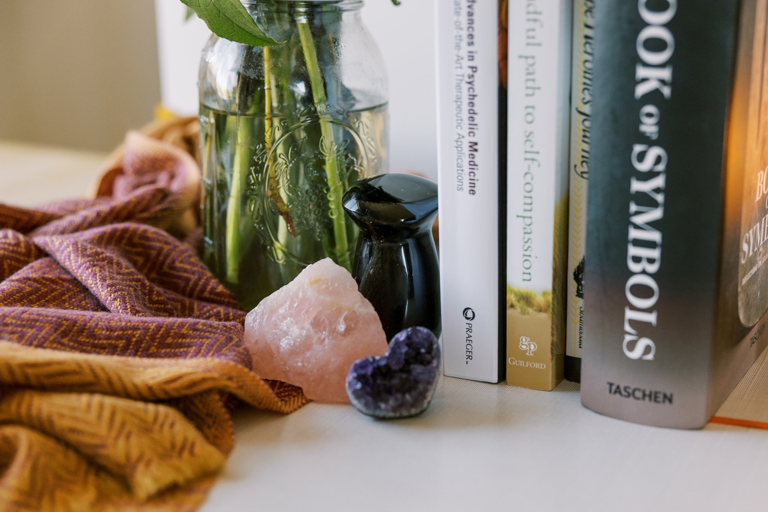 A glass jar with green leaves, a pink Himalayan salt rock, a small purple amethyst, a black essential oil diffuser, and a row of books on topics such as medicine and subconsciousness.