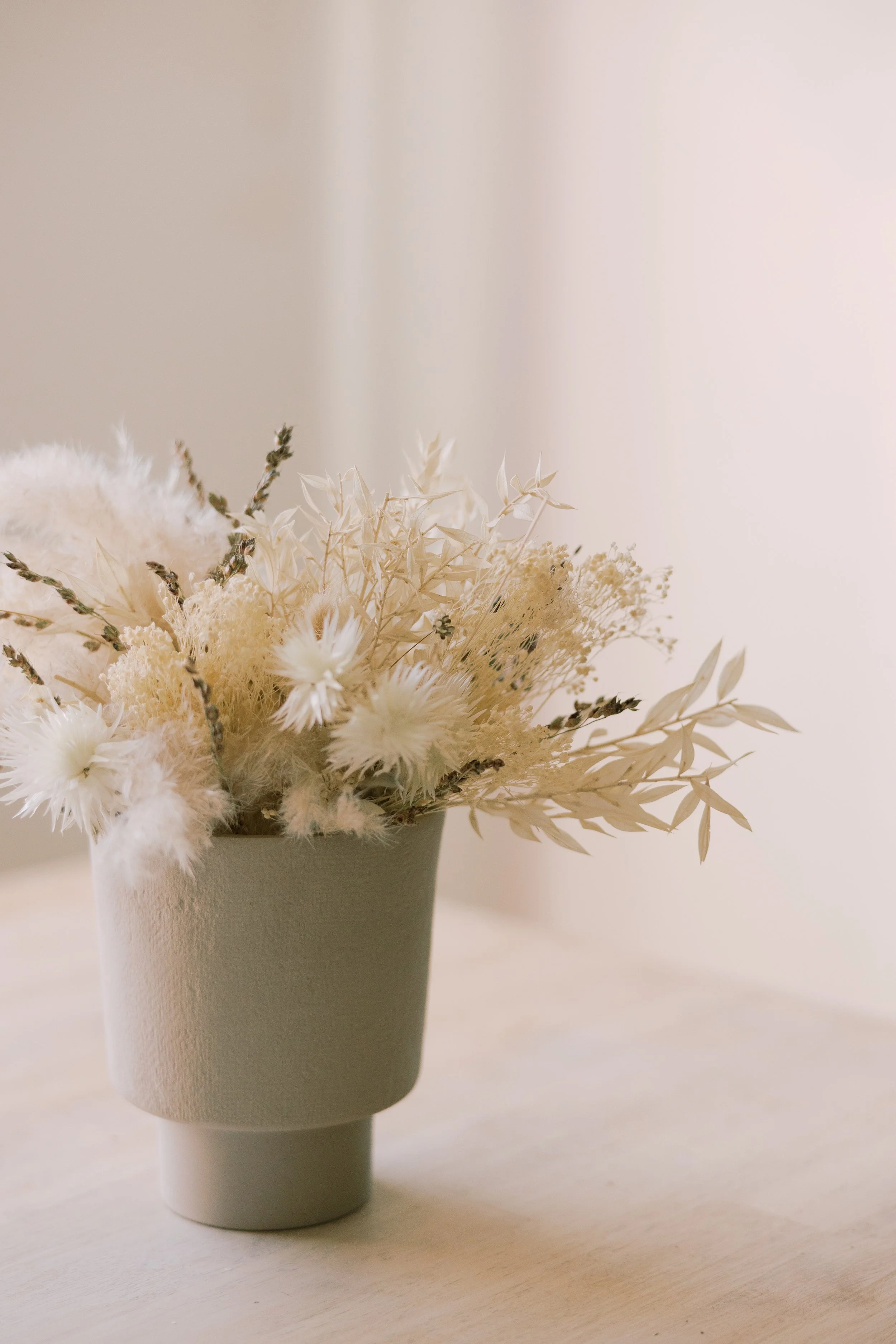 A beige vase with dried white and cream-colored flowers and foliage on a light wooden surface against a soft off-white background.