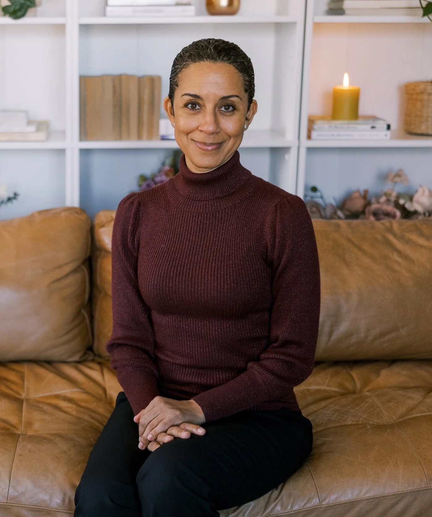 A woman with dark curly hair pulled back, wearing a maroon turtleneck sweater, sitting on a tan leather couch in a cozy room with bookshelves, a lit candle, and decorative items in the background.