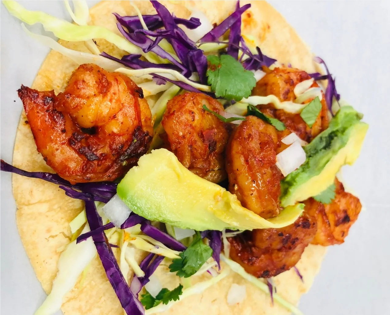 Spicy shrimp Taco with shredded purple cabbage, sliced avocado, chopped onions, cilantro, and shredded lettuce in a corn tortilla.