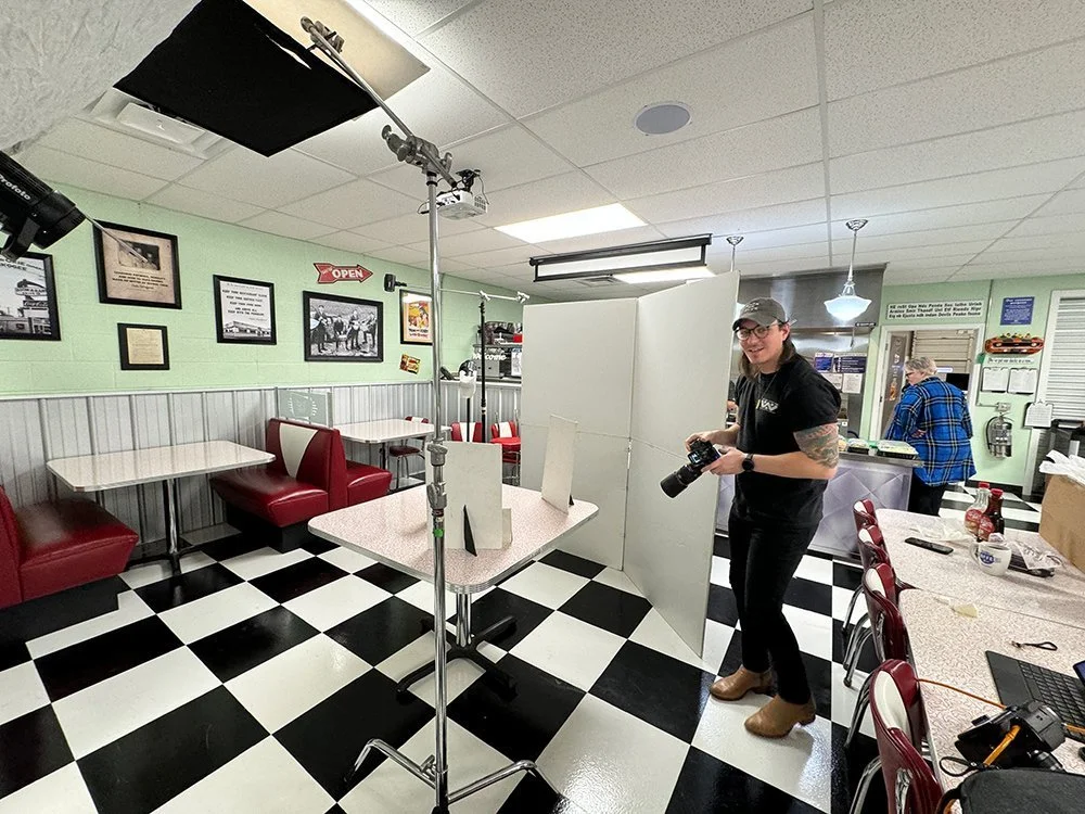A woman holding a camera stands behind a white room divider inside a diner with checkered black and white floor tiles, red booths, and wall decorations.