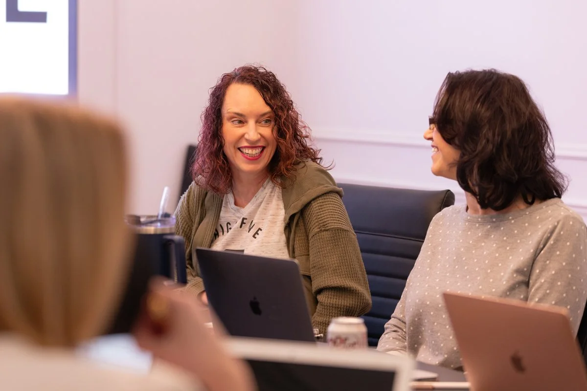 Two women sitting at a table, smiling and talking in a meeting room, with laptops and drinks in front of them.