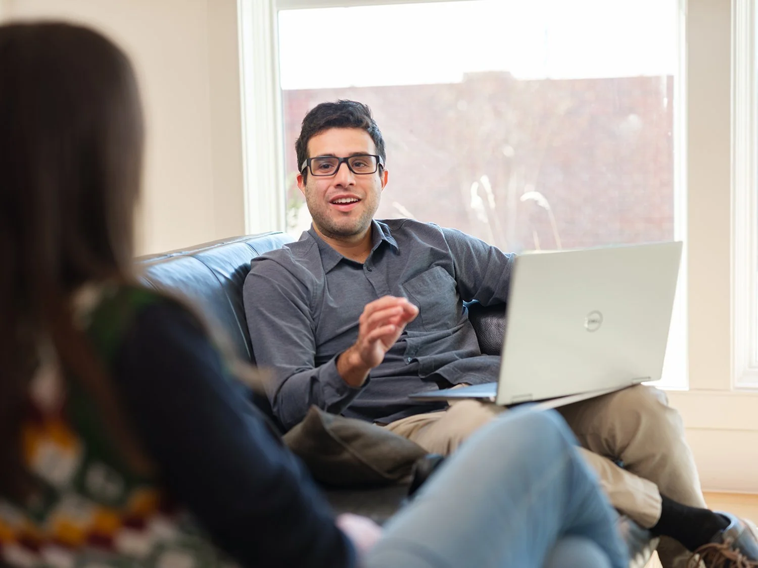 A man wearing glasses and a gray shirt sitting on a sofa with a laptop, talking to a woman in a patterned shirt sitting across from him, in a room with large windows and natural light.