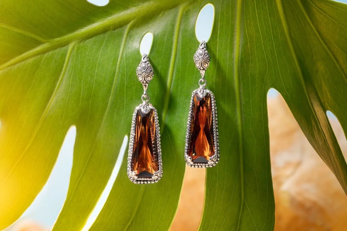 A pair of dangling earrings with rectangular orange gemstones and silver decorative details, hanging from a green tropical leaf.