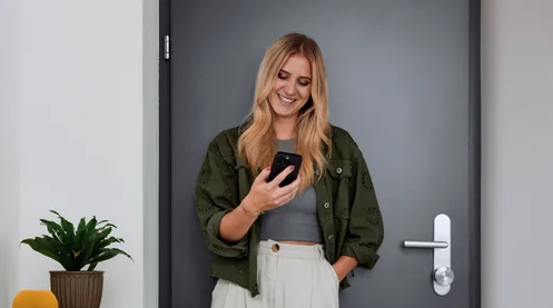 A smiling woman with blonde hair looking at her phone in front of a gray door, with a small potted plant on a table to her left.