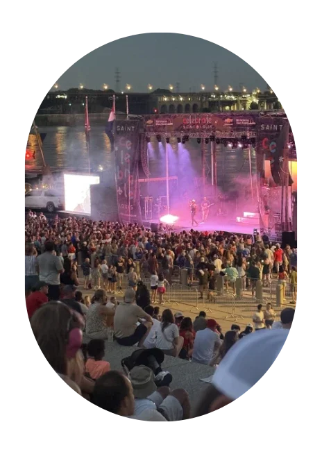 Outdoor music concert on a riverbank during evening with a large crowd watching performers on a stage illuminated with purple lights.