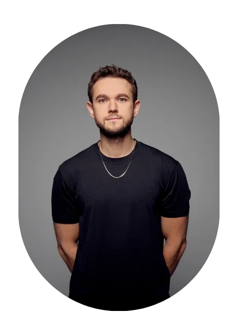 A young man with short brown hair and a beard standing against a gray background, wearing a black t-shirt and a silver chain necklace.