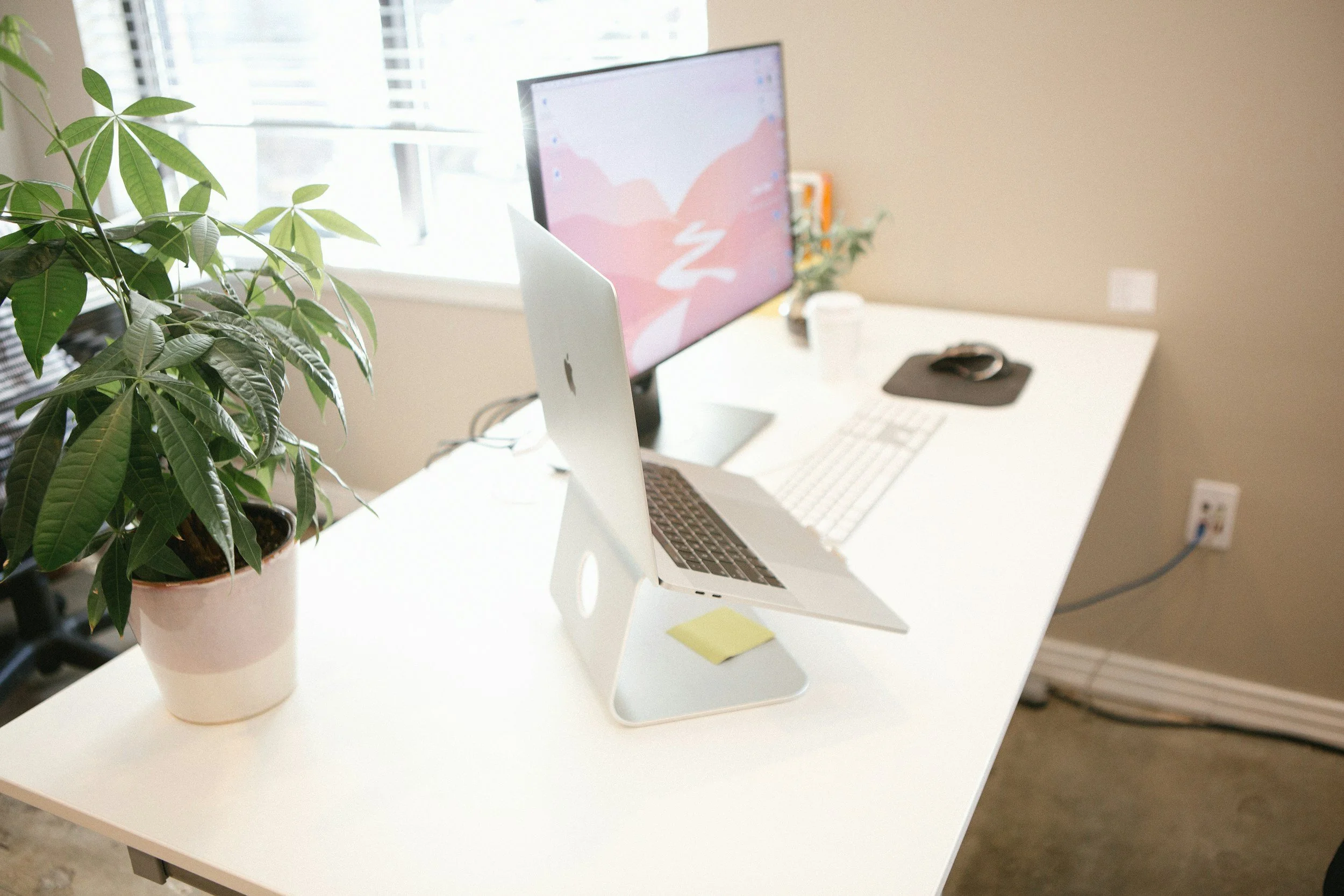 A white desk with a potted plant, an Apple computer monitor, a white keyboard, a black mouse on a mouse pad, and sticky notes, near a window with blinds in a well-lit room.