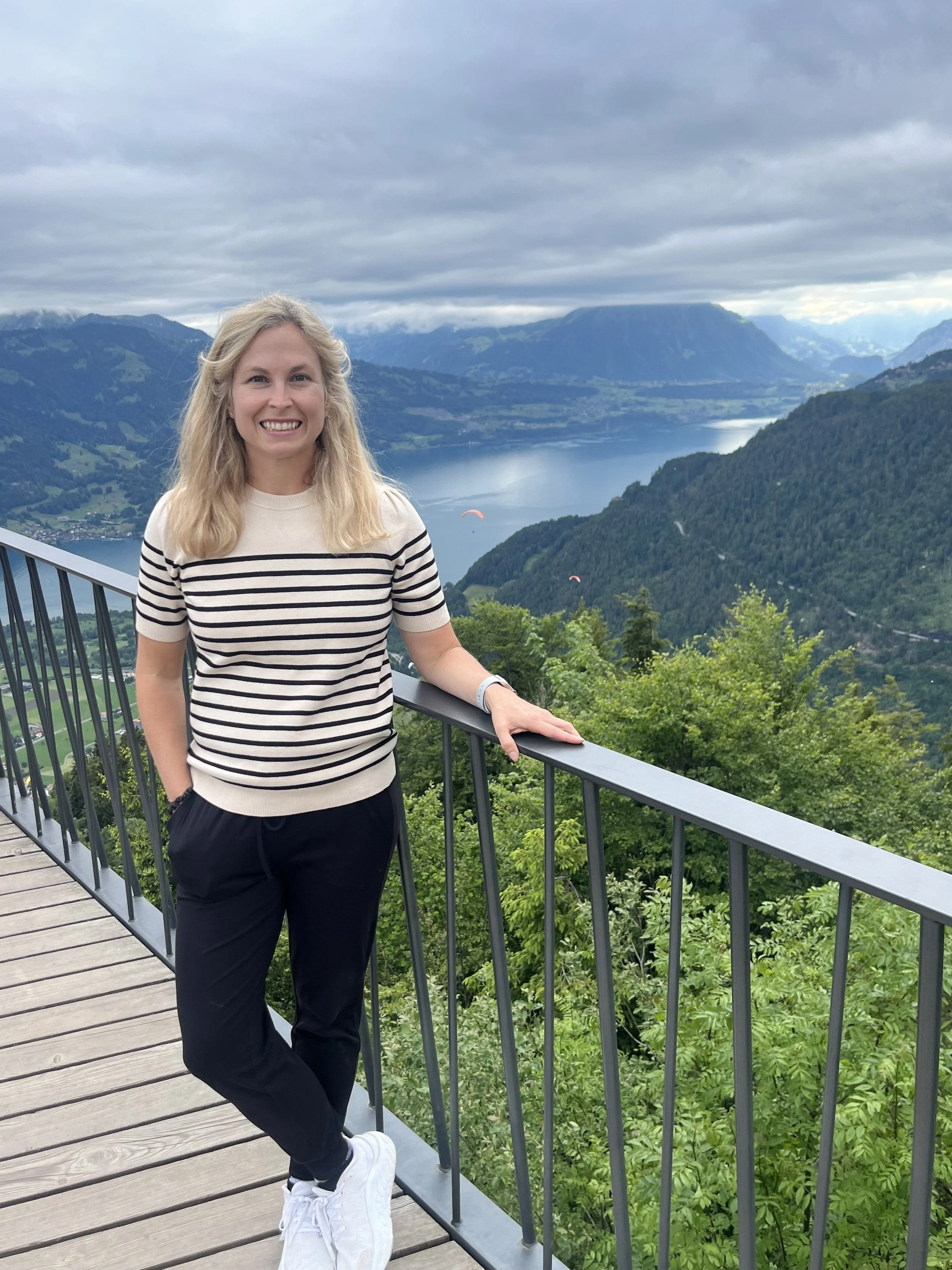 A woman with blonde hair, wearing a striped shirt, black pants, and white shoes, standing on a viewing platform with a scenic background of a lake, green mountains, and a cloudy sky.