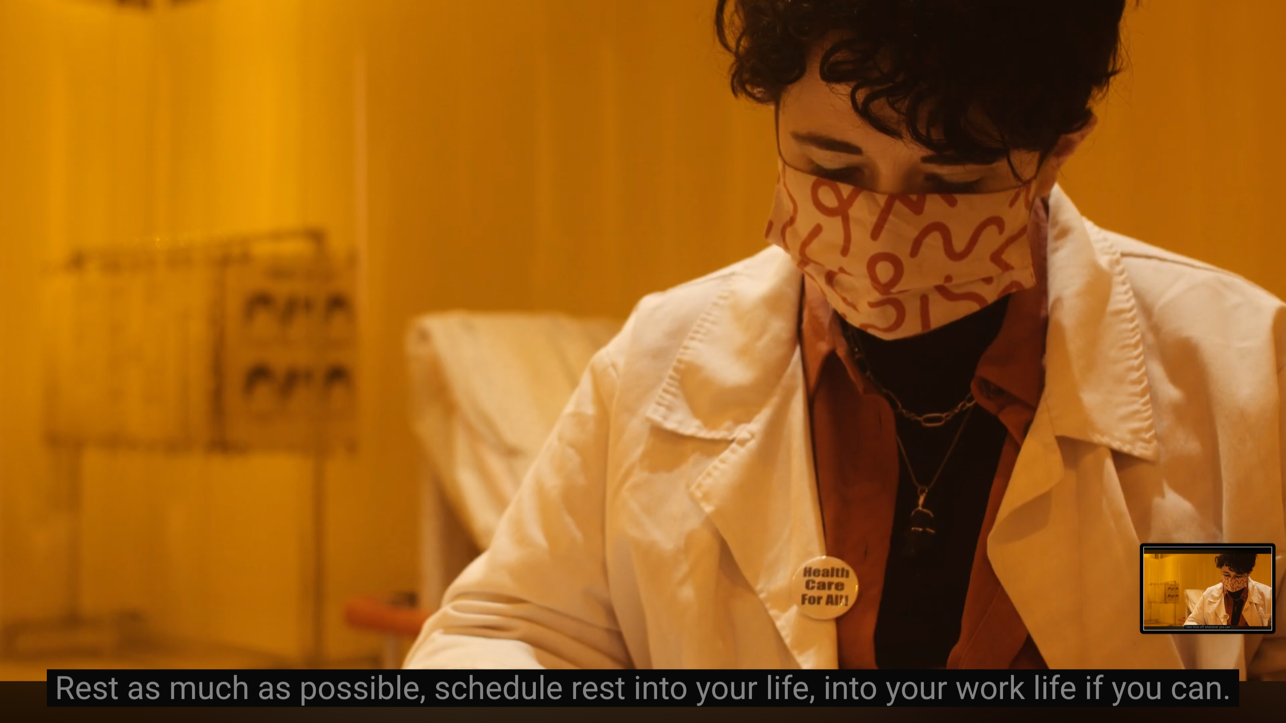 Close up of Aine a white person with short black curly hair, they are wearing a pink surgical mask and a white lab coat along with a badge that says 'healthcare for all'. They are sitting within their yellow partially see through hospital style curta