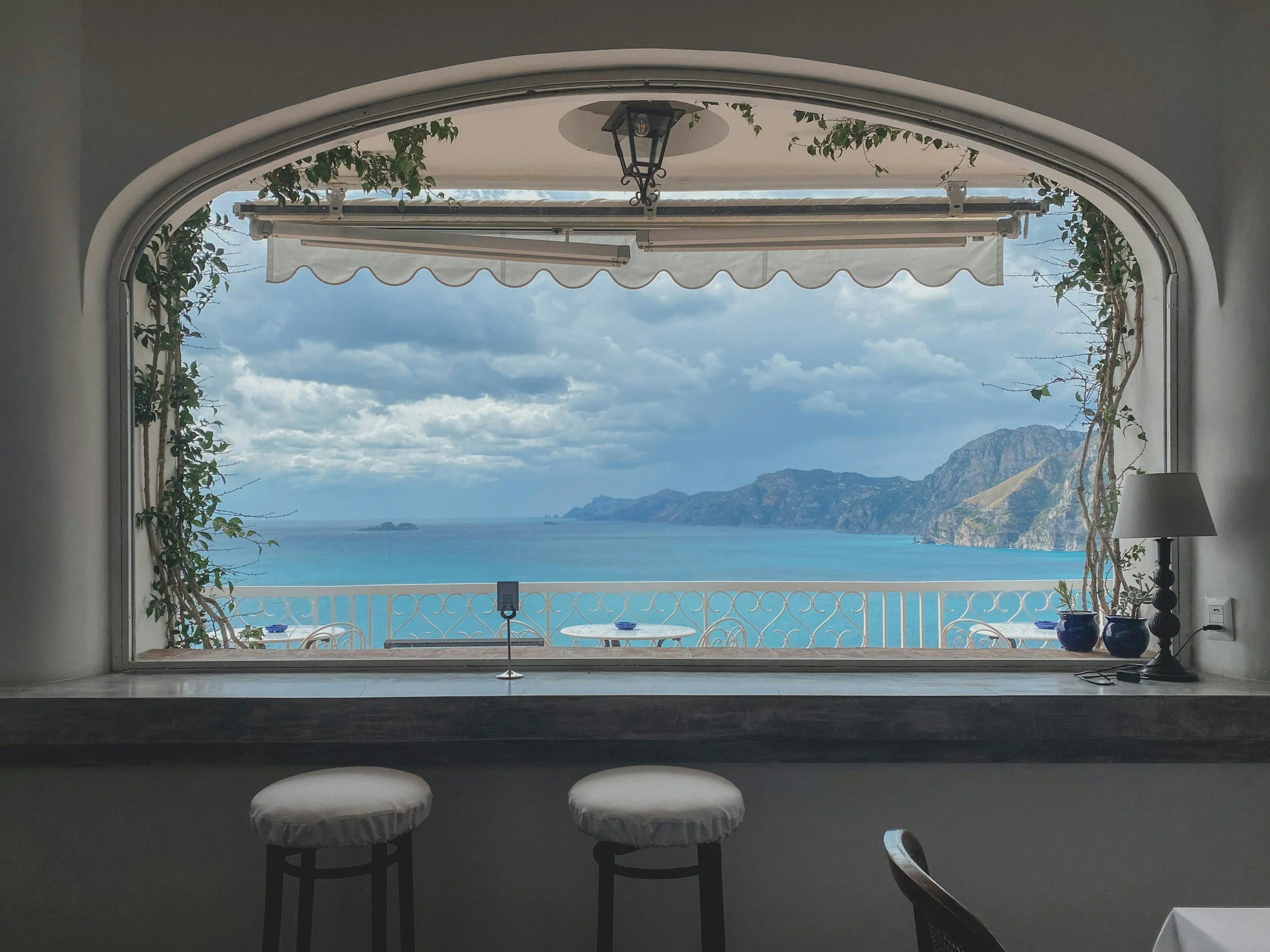 View of the ocean and distant mountains through a large arched window, with plants on the sides and a balcony with a white iron railing outside.