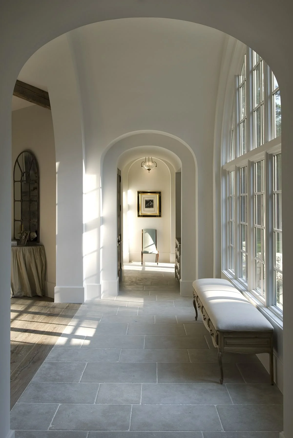 Bright hallway with large windows, stone tile floor, and arched ceiling, featuring a bench by the window and artwork at the end.