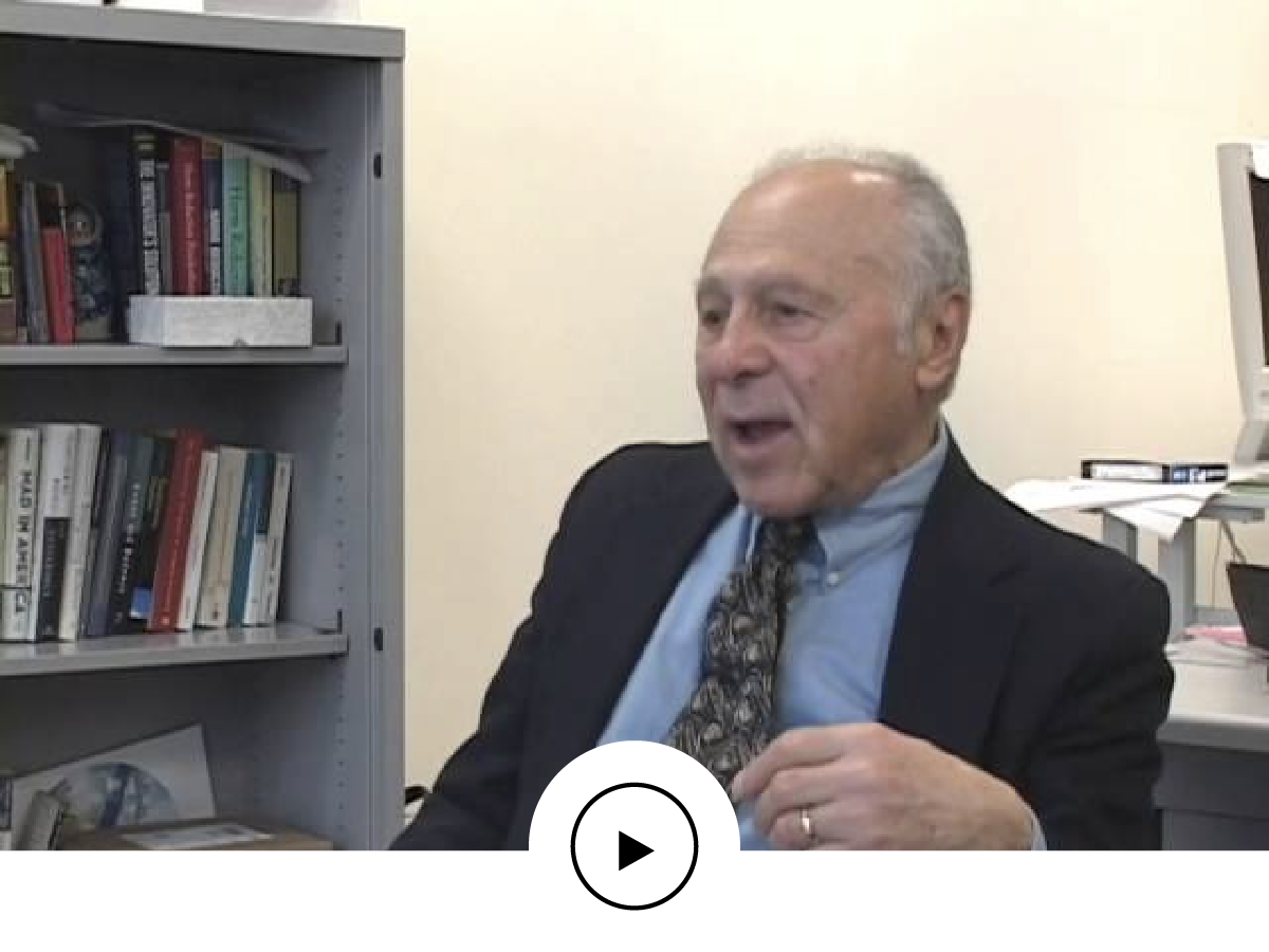 An older man in a suit and tie sitting in an office, speaking, with a bookshelf full of books on his left and a computer on his right.