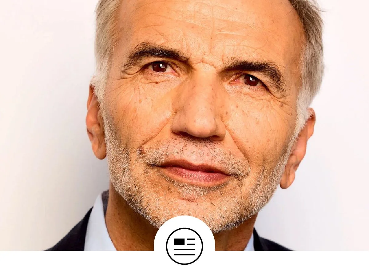 Close-up portrait of an older man with gray hair and a beard, wearing a suit and tie, smiling gently, against a plain background.