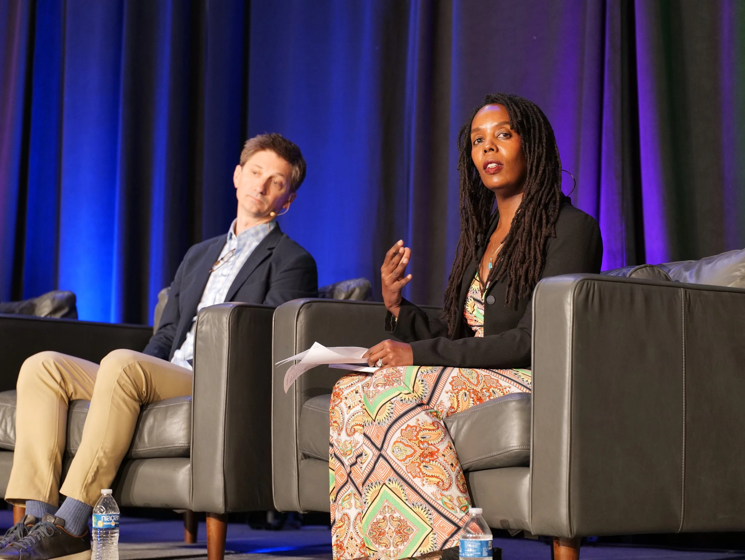 A woman giving a presentation at a conference, standing at a podium with a laptop, with a large screen behind her displaying slides about PSH site-based projects and illustrations of housing developments, while an audience of diverse people listens attentively.