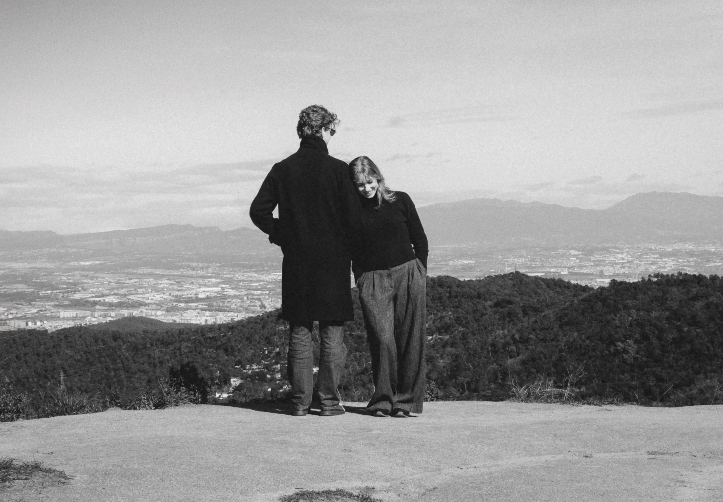 Twee mensen staan samen op een bergtop, omgeven door een uitgestrekt landschap met bergen in de verte, in zwart-wit gefotografeerd.