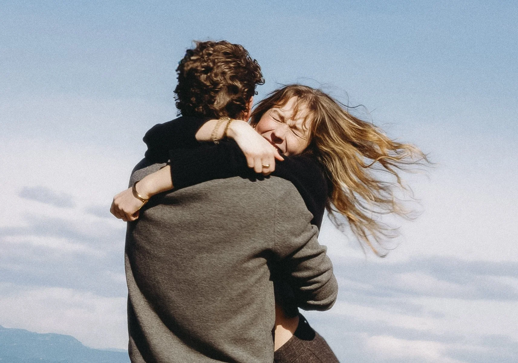 Twee mensen die elkaar omhelzen en lachen, onder een blauwe lucht met wolken. couple photography, travel documentation, travel photography, Belgian photographer