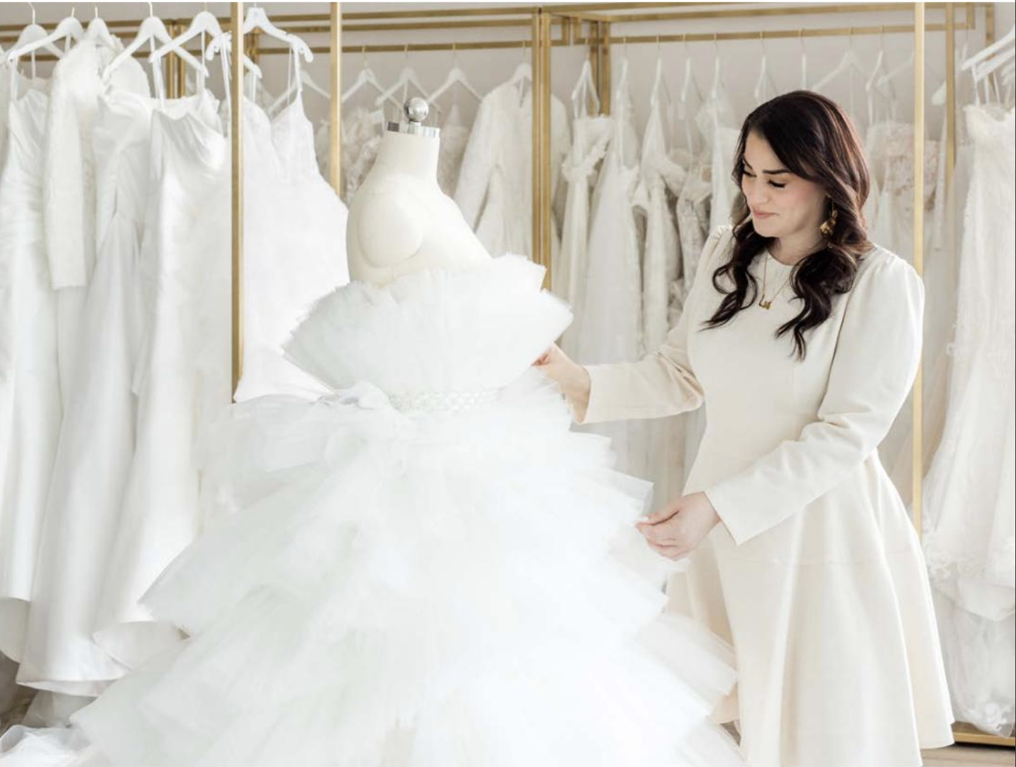A woman in a cream-colored dress looking at a white wedding gown on a dress form, surrounded by other white wedding dresses in a boutique