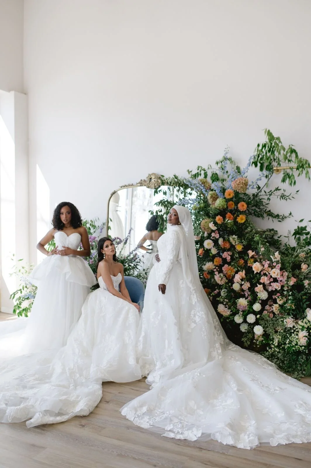 Three women in wedding dresses in front of a large floral arrangement and mirror, with natural light coming from the side.