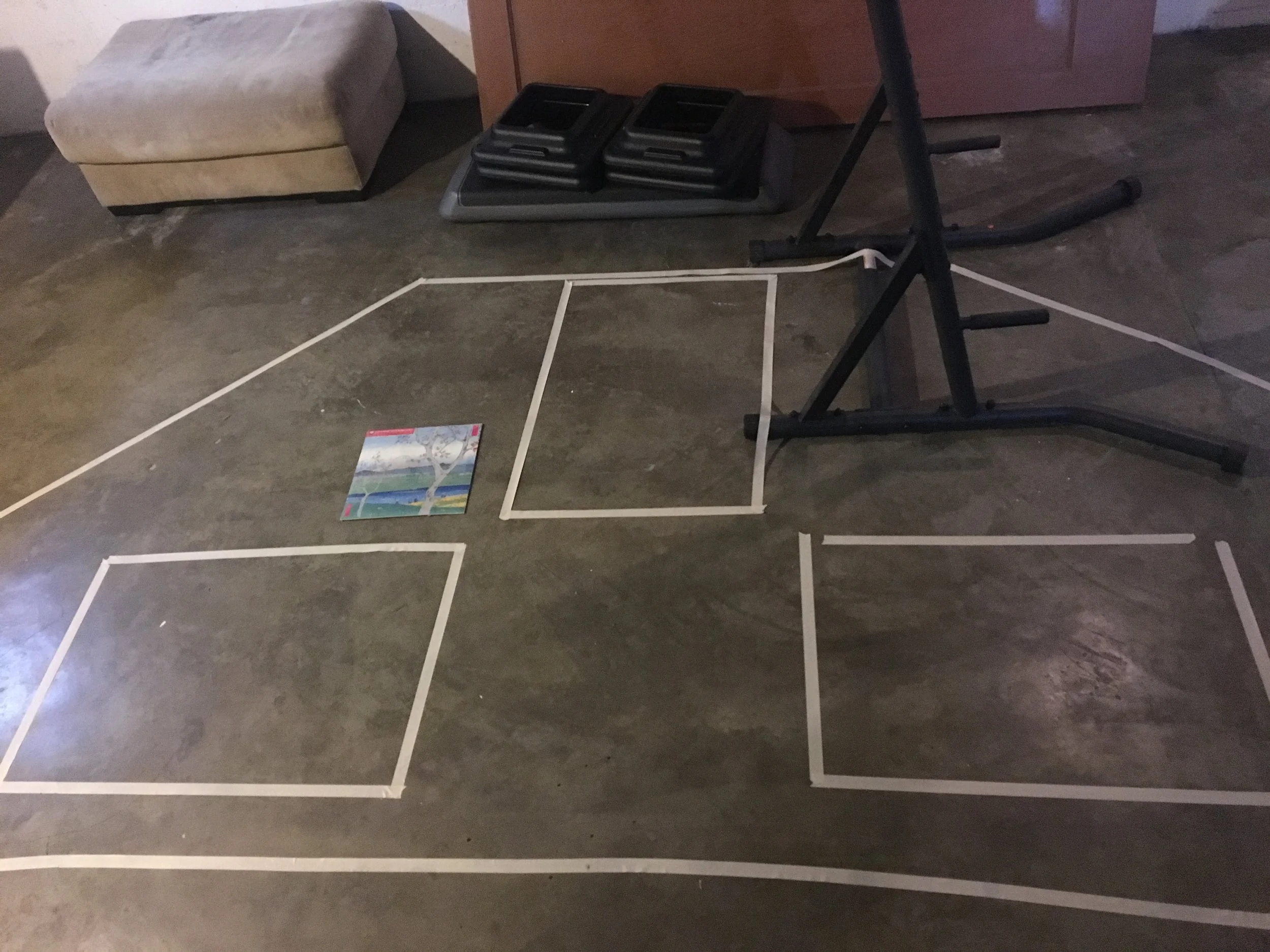 Masking tape along a cement floor marks the outline of one of the slanted walls from the room, with the outlines of big posters taped within the shape, to visually see how many posters would fit on the wall.