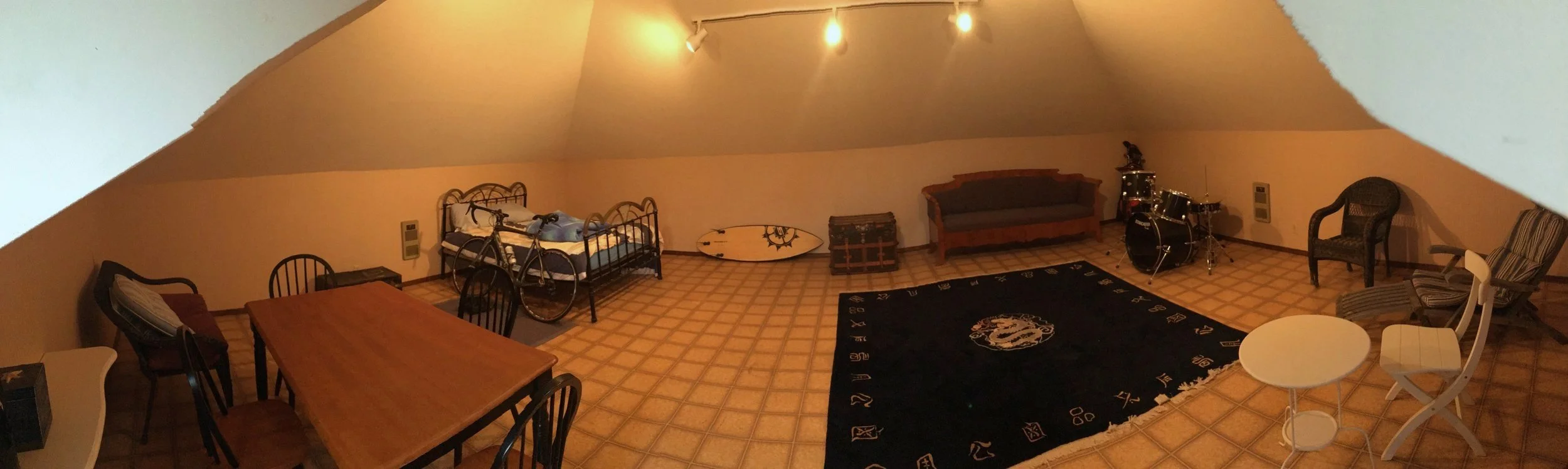 Far left corner has a metal bed frame, with blue bedding, and moving left to right along the wall there is a surfboard, an old wooden chest, and a church pew.