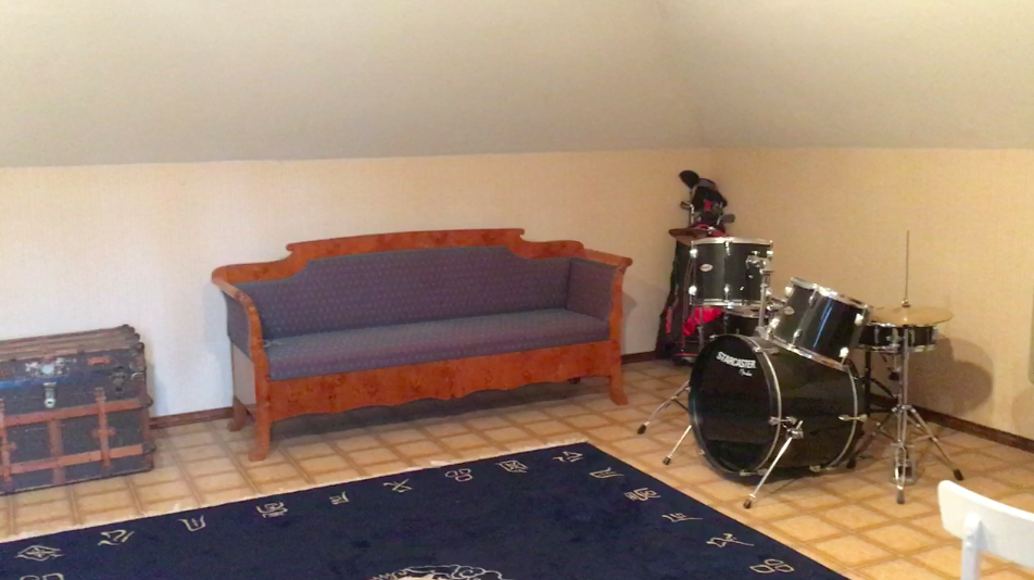 In the right corner of the room sits the church pew - with an amber color wood stain and blue dot patterned upholstered cushions - a set of golf clubs and complete black drum set.