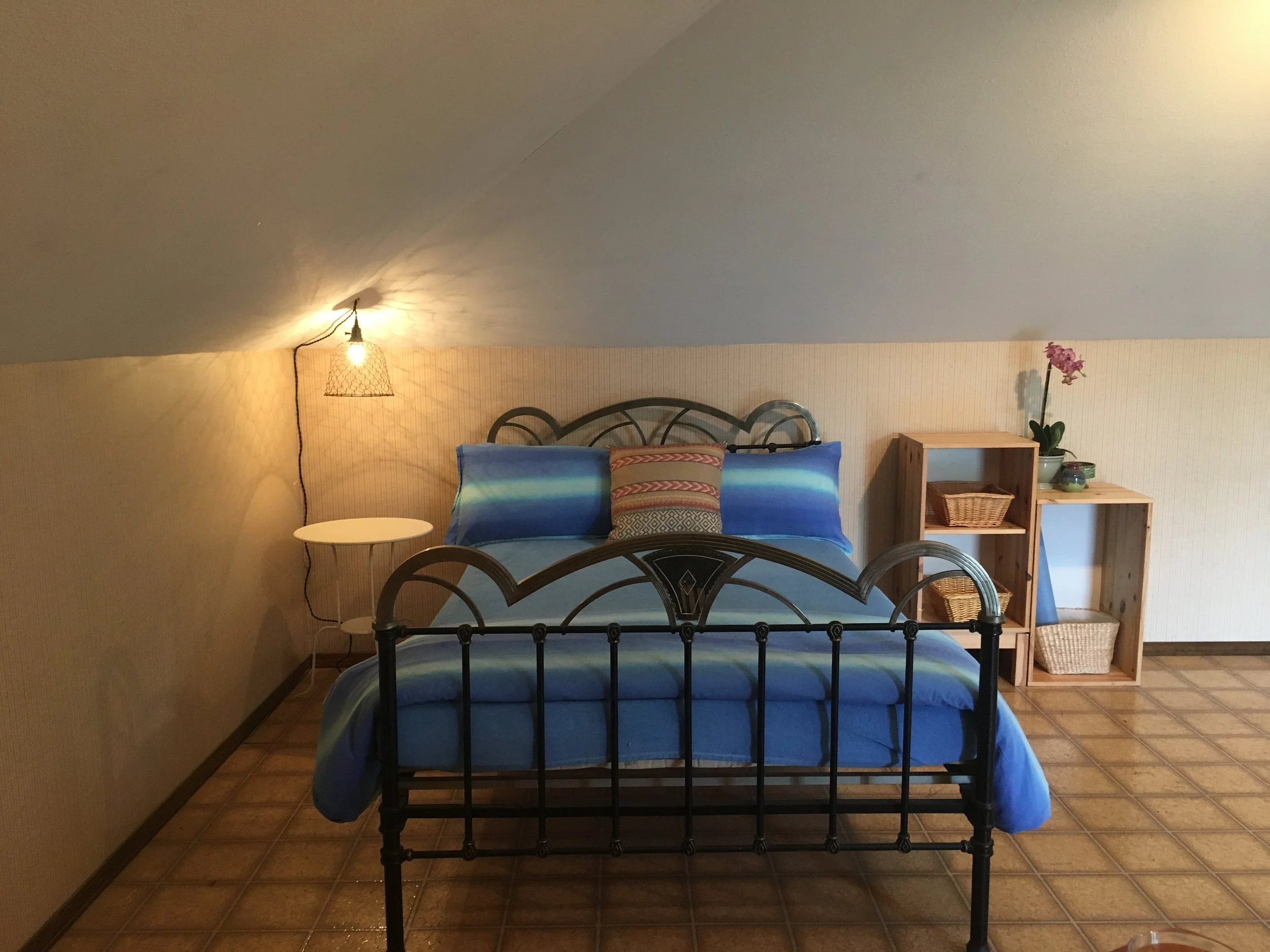 Antique Art Deco metal bed frame with blue bedding. A white night stand table is on the left wit a pendant light suspended above. To the right is exposed wood-grain open storage for some wicker storage baskets and a blue yoga mat.