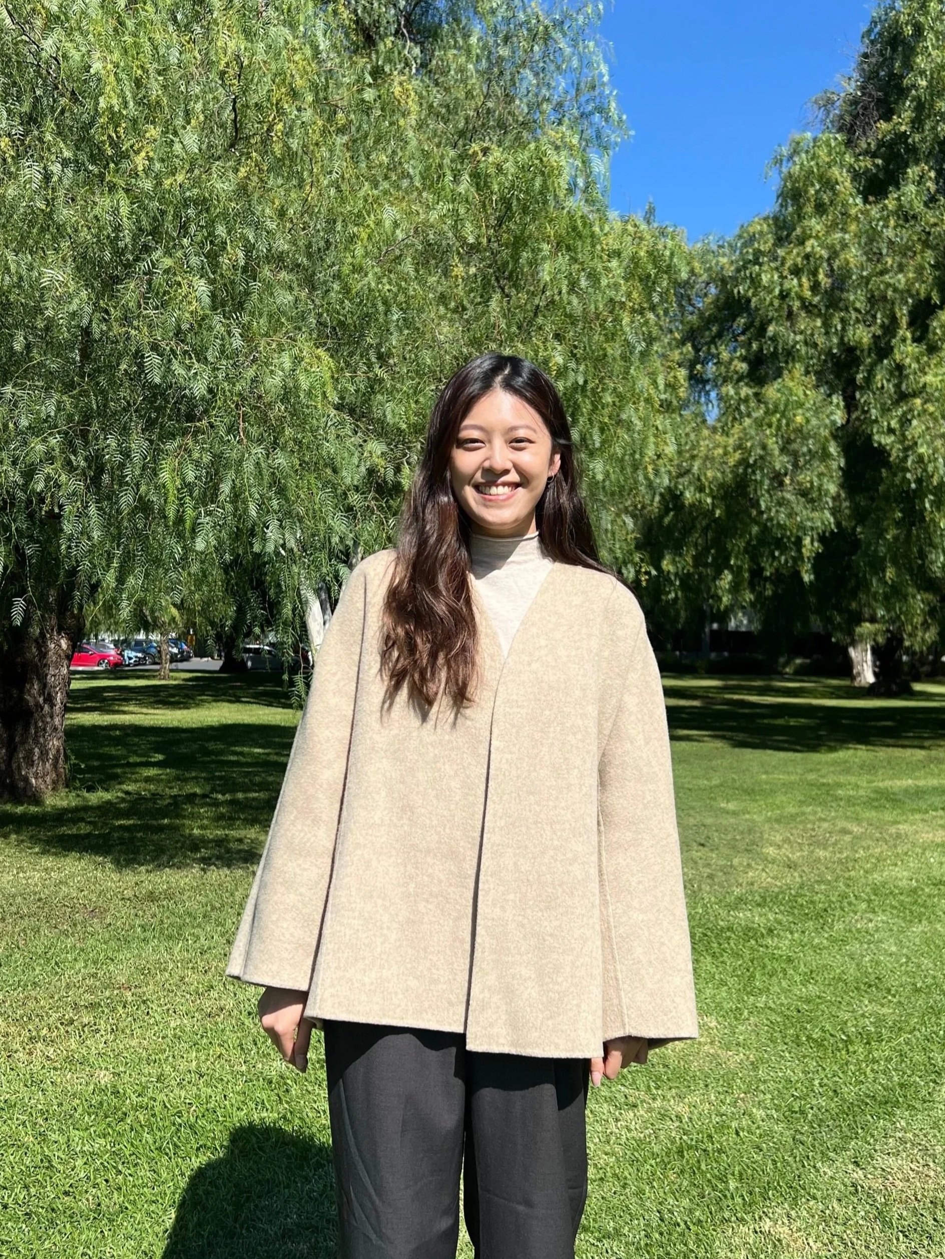 A young woman with long dark hair smiling outdoors on a sunny day, standing on grass with trees and a blue sky in the background.