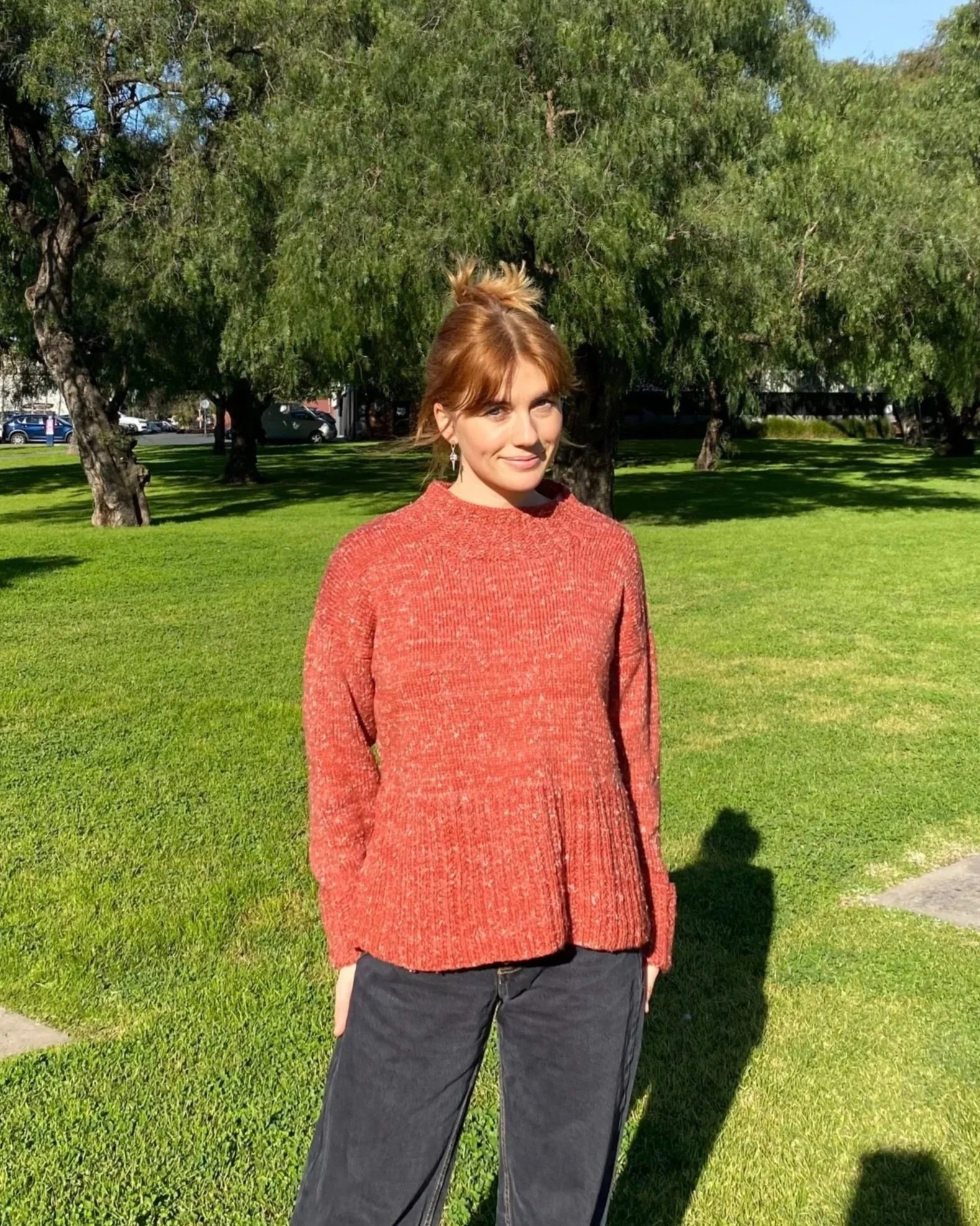 A woman with red hair tied in a bun wearing a red sweater and black pants stands on a grassy field with trees in the background.