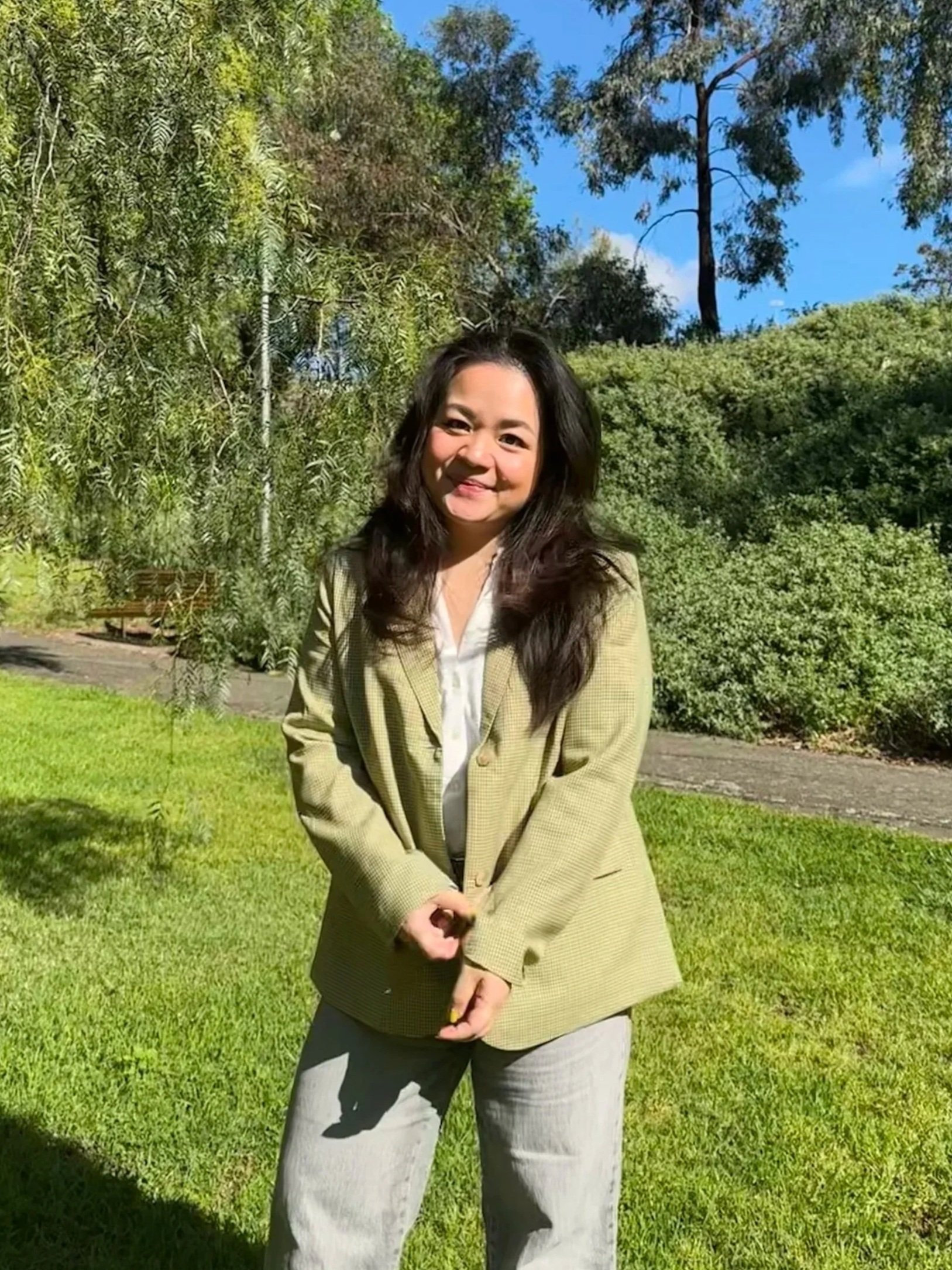A woman standing outdoors on a sunny day with lush green trees and grass in the background.