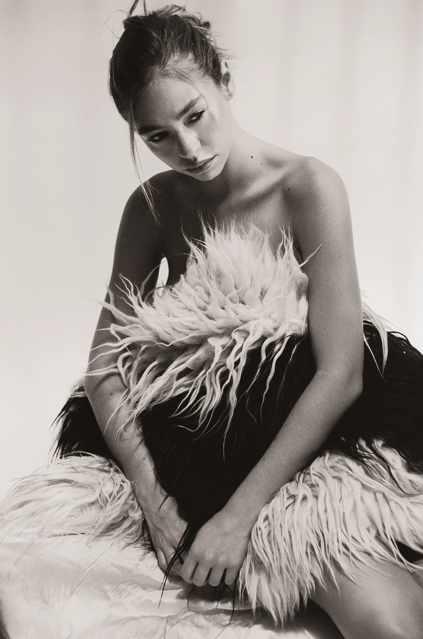 A woman with dark hair in a messy bun, wearing a strapless dress with feather details, sitting on a fluffy feathered rug, looking downward with a serious expression.