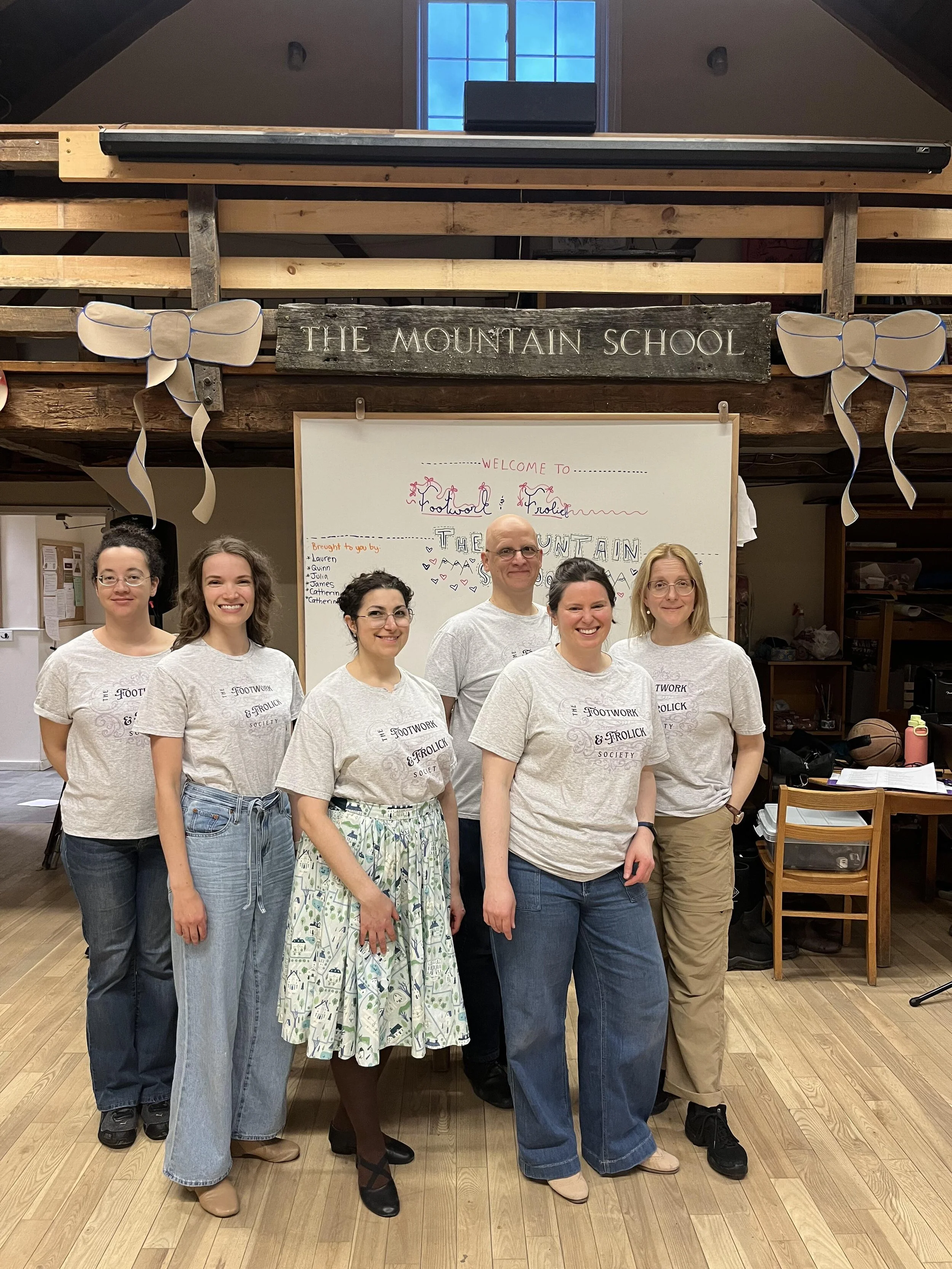 a ground in Footwork and Frolick t-shirts smile at the camera with a sign reading The Mountain School visible above their heads