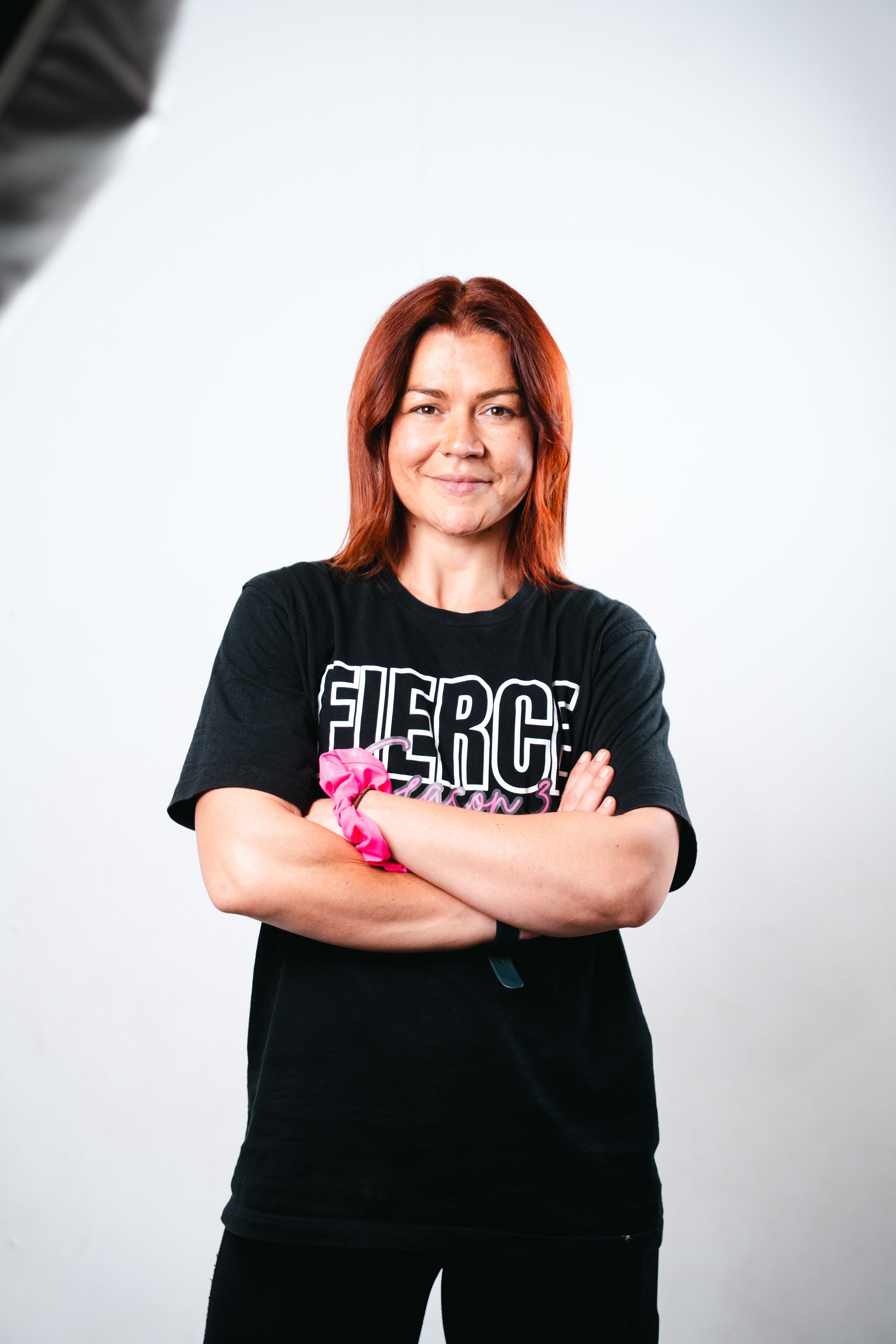 Woman with short brown hair smiling and standing with arms crossed, wearing a black T-shirt with the word 'FIERCE' on it, against a white background.