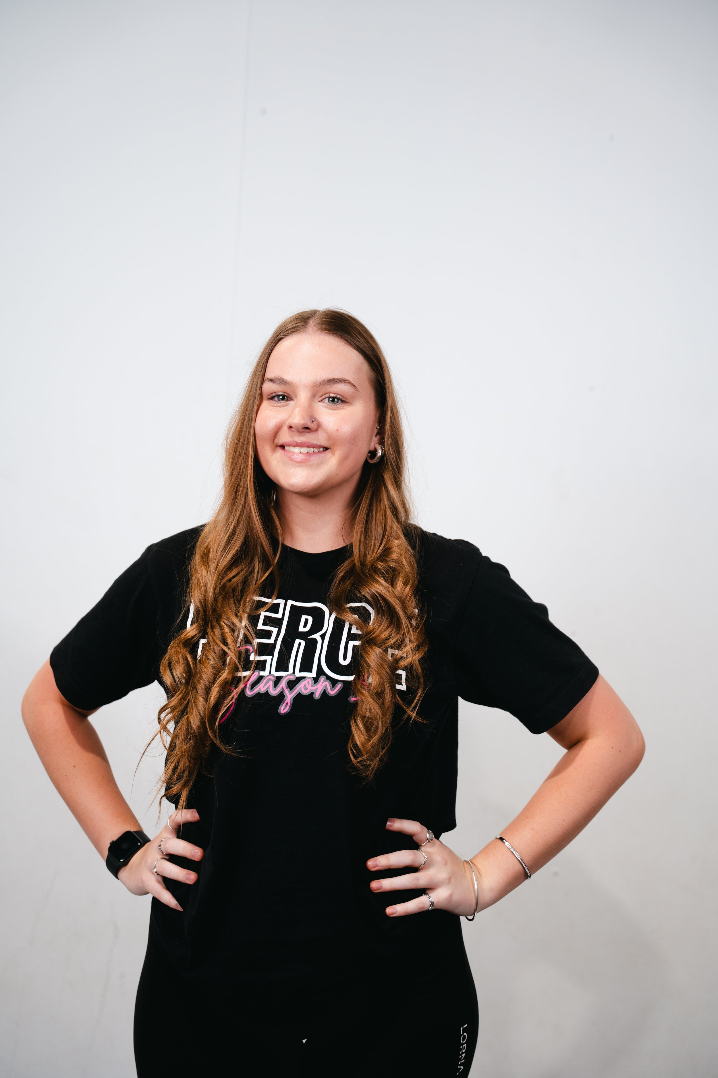 A young woman with long straight hair, wearing a black T-shirt that says 'PERFORMANCE ARTS' inside a decorative design, standing with hands on her hips and smiling, against a plain white background.