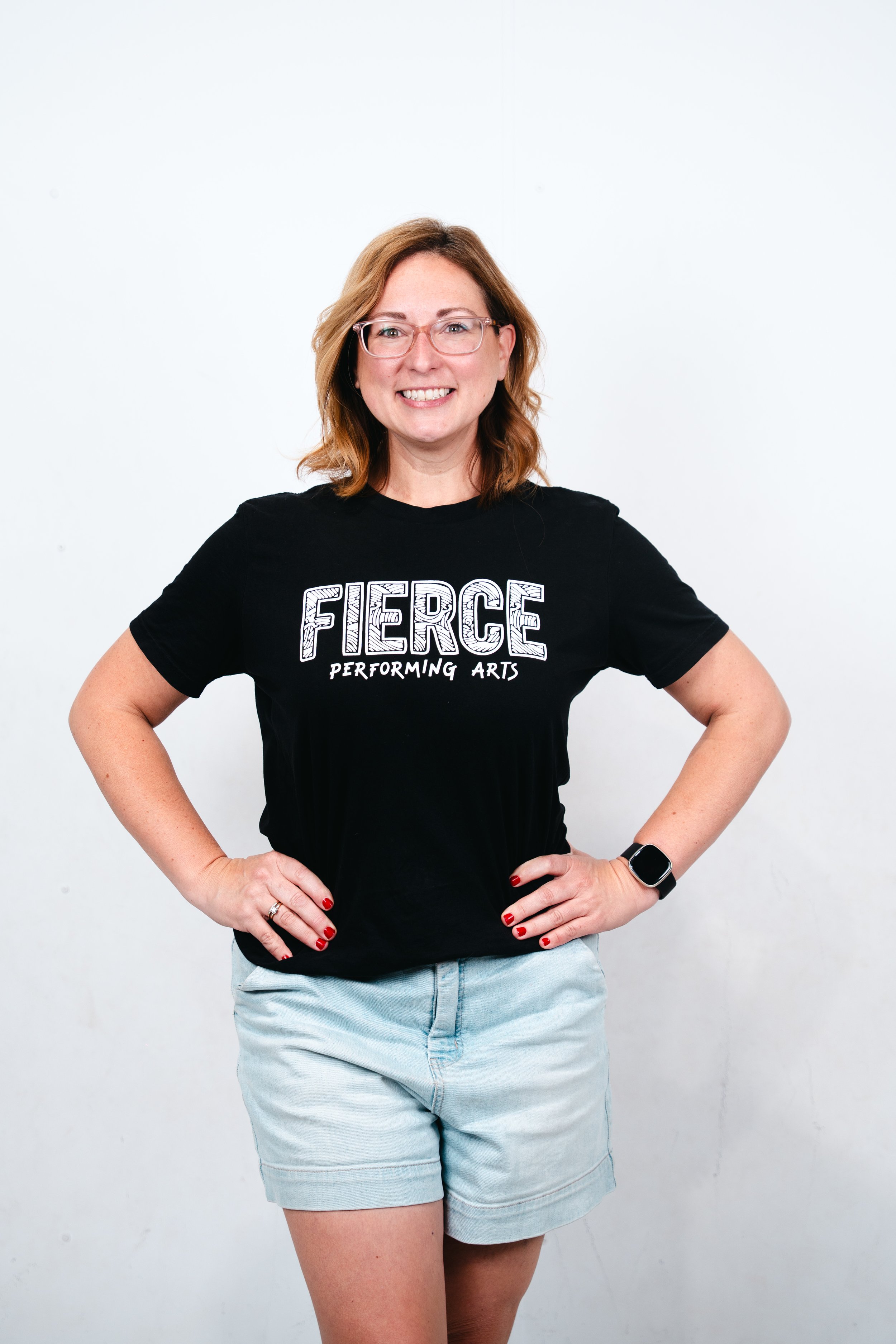 Woman with shoulder-length auburn hair smiling, wearing a black T-shirt that reads 'FIERCE PERFORMING ARTS,' and standing with her hands on her hips against a plain white background.