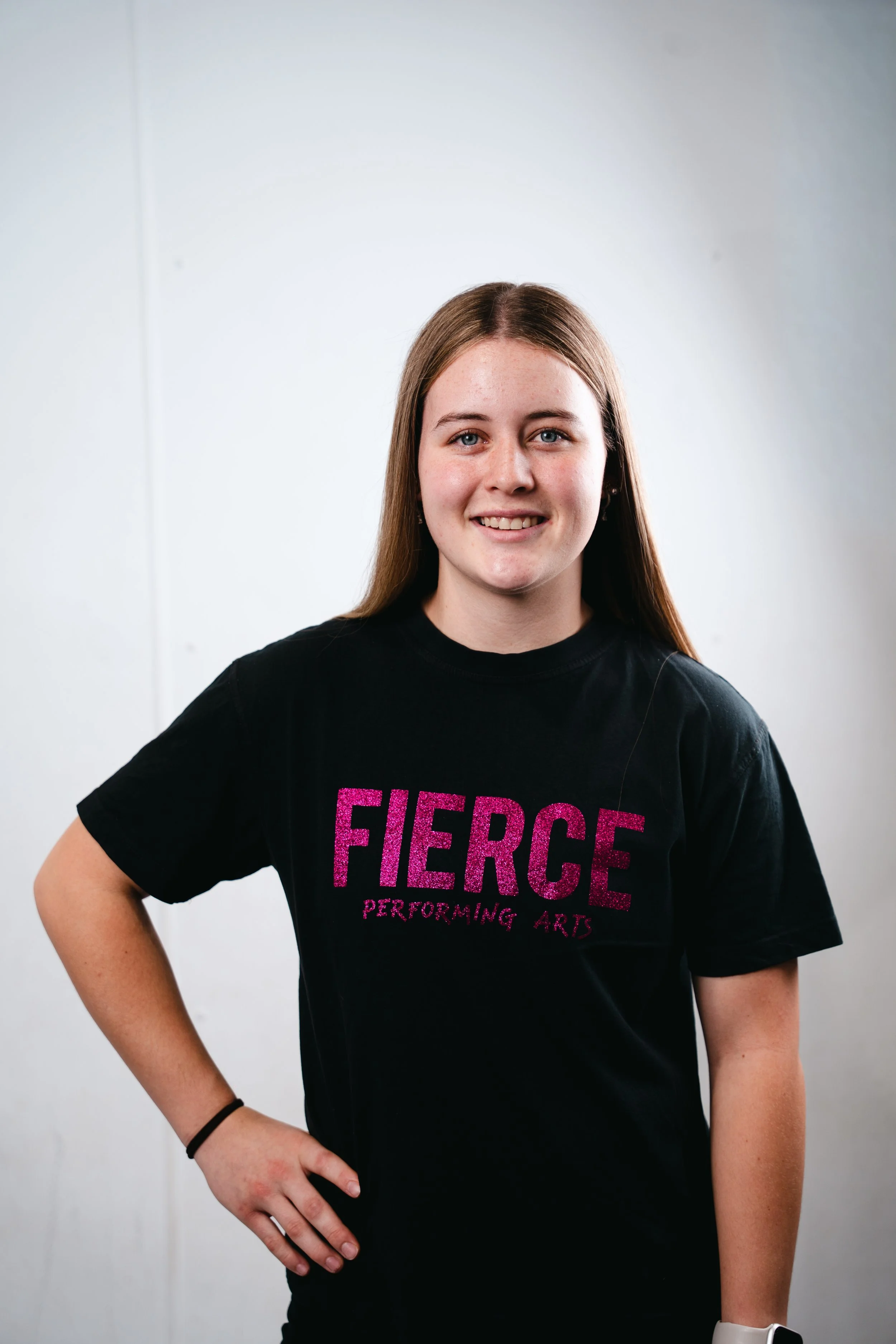 A smiling woman with long black hair, wearing a black t-shirt with pink text that says 'FIERCE Performing', stands with one hand on her hip against a plain white background.