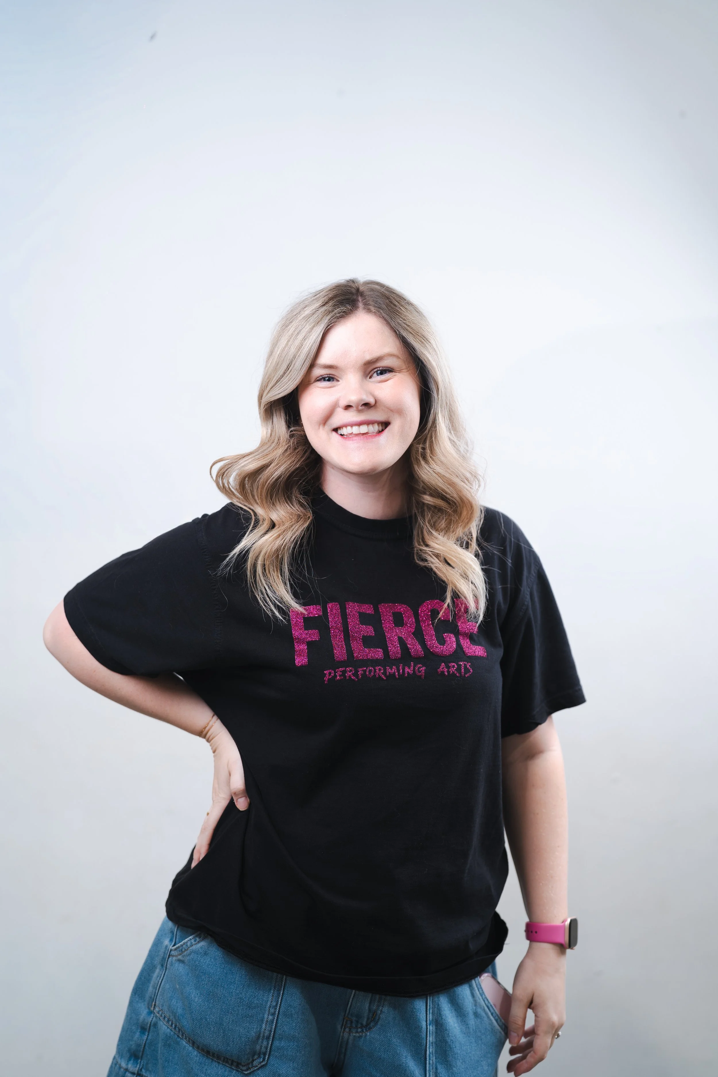 A woman with shoulder-length curly hair wearing a black t-shirt with purple text, smiling and posing with one hand on her hip, against a plain white background.