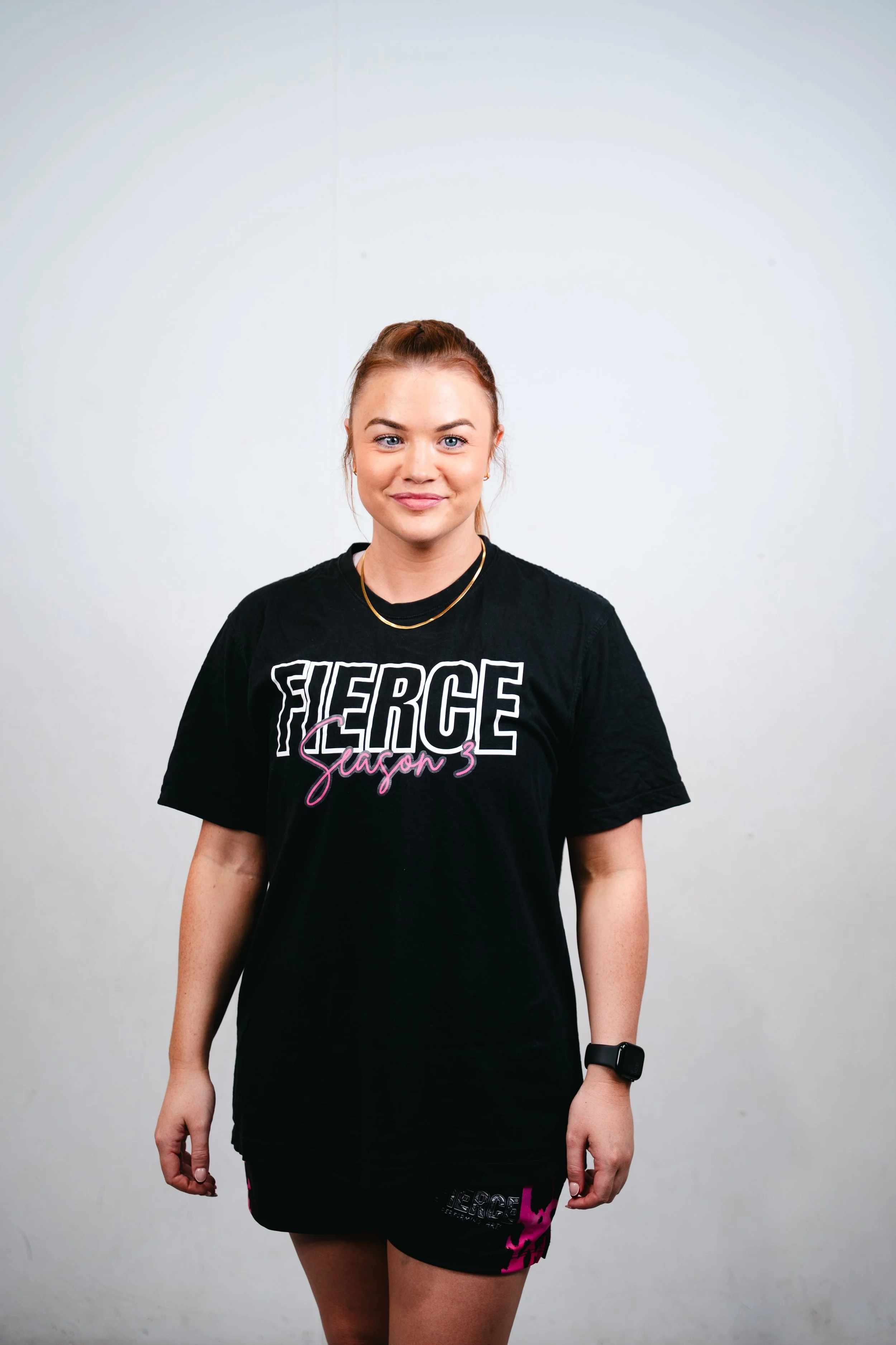 A young woman with shoulder-length brown hair, smiling, wearing a black t-shirt with 'FIERCE Performing Arts' printed on it, standing against a plain white background.