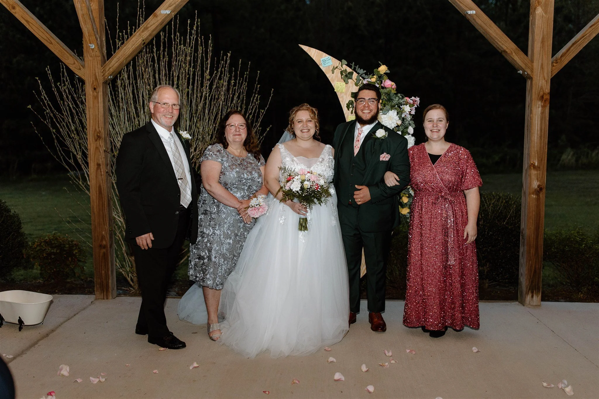 A wedding group photo featuring five people, including a bride in a white gown holding a bouquet, a groom in a dark suit, and three other family members, outdoors at night with a wooden arch and floral decorations.