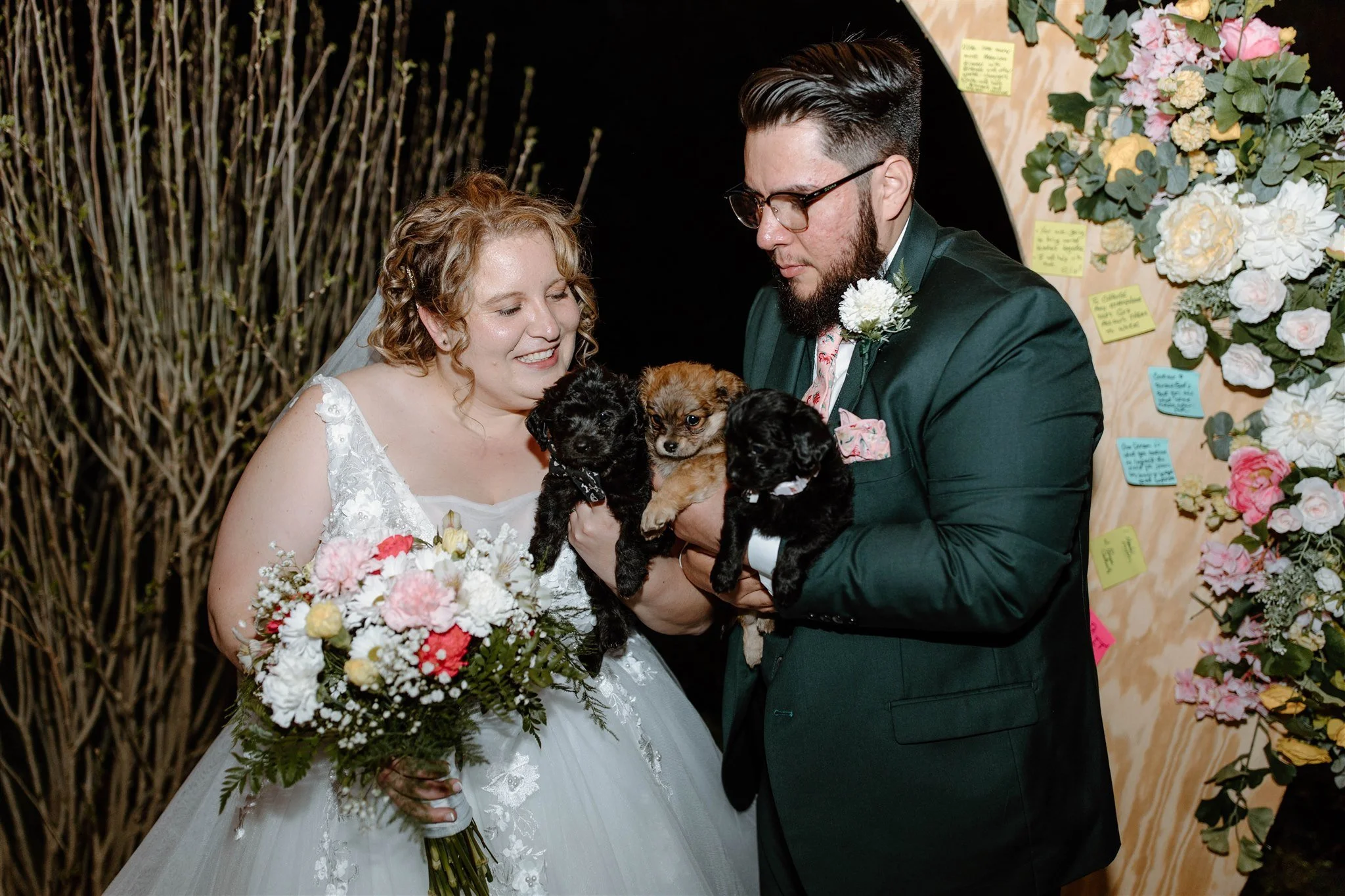 A bride and groom holding puppies during their wedding. The bride is wearing a white dress and holding a bouquet of flowers. The groom is wearing a dark green suit and glasses. They are smiling and looking at the puppies they are holding. The backgro