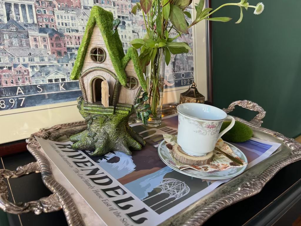 Decorative tray with a cup and saucer, a small spoon, a green moss-covered fairy house, a vase with green leaves and white flowers, a magazine titled 'ABANDONED,' and a framed art piece of buildings in the background.