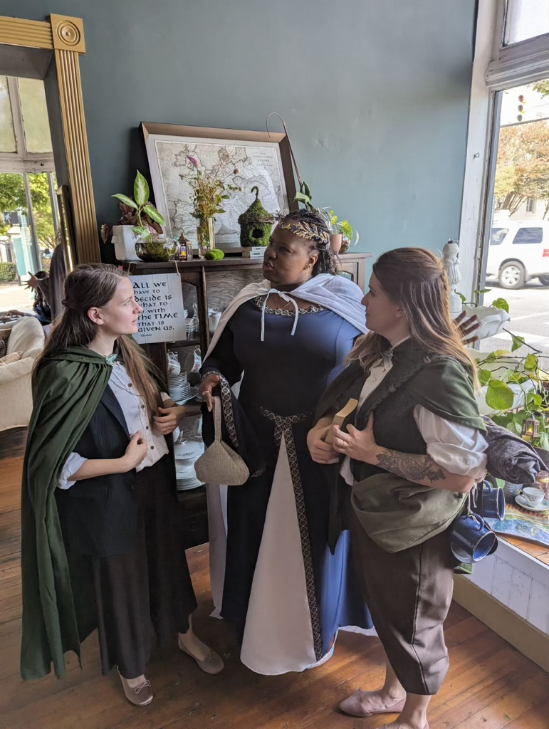 Three women, dressed in historical or fantasy costumes, are having a conversation inside a cozy room with a large window. The woman in the center is wearing a black dress with a white cape and a laurel wreath crown, while the other two women are wear