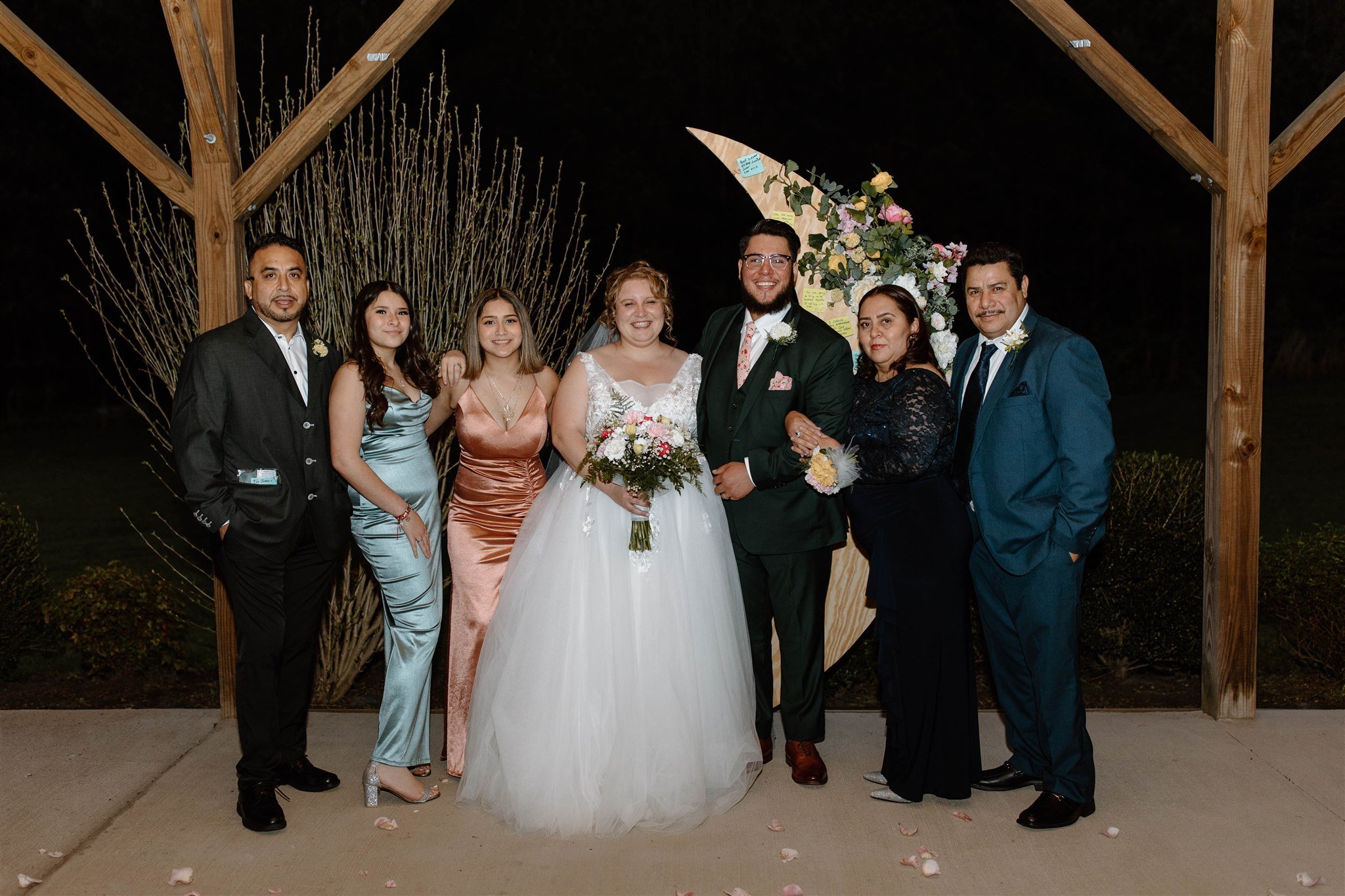 Group of people at a wedding celebration, standing outdoors at night under a wooden structure with floral decorations, including the bride and groom in the center, surrounded by family and friends.