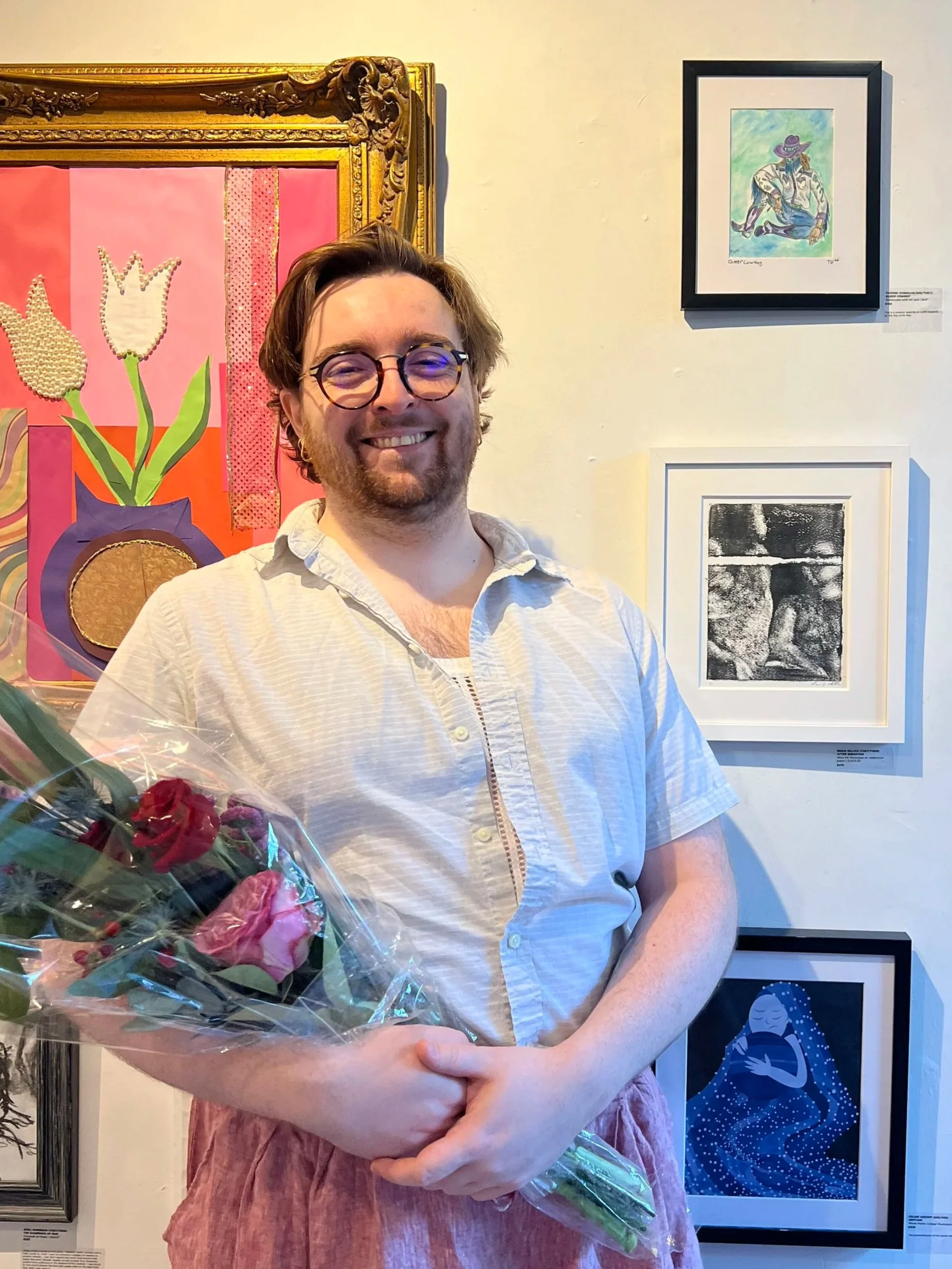 A man with glasses smiling and holding a bouquet of pink and red roses, standing in front of framed artwork on a gallery wall.