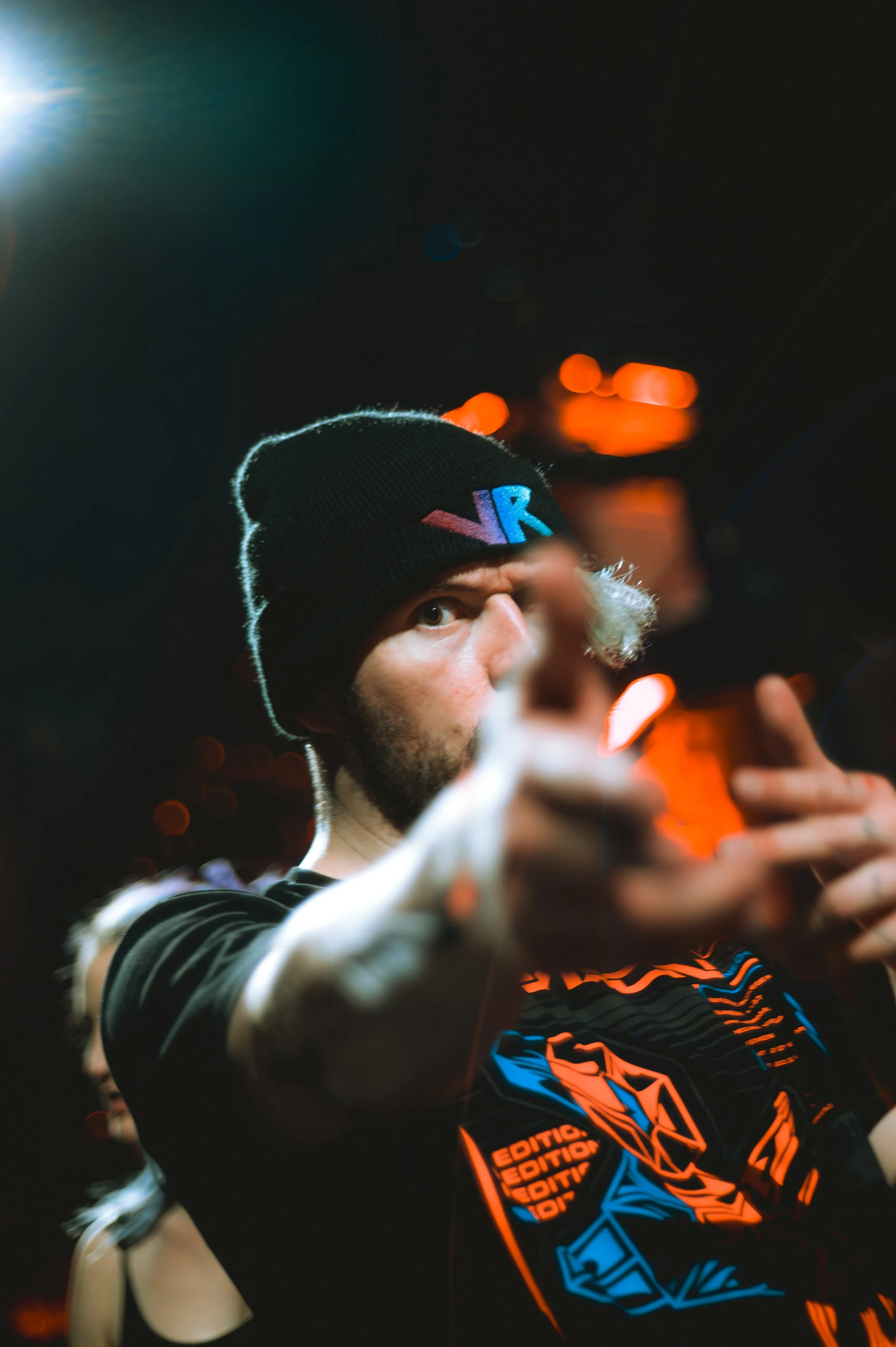 Close-up of Virtual Riot wearing a black VR beanie and a graphic t-shirt. He points toward the camera with a blurred hand in the foreground. Intense stage lighting creates sharp highlights and warm orange bokeh in a moody concert setting.