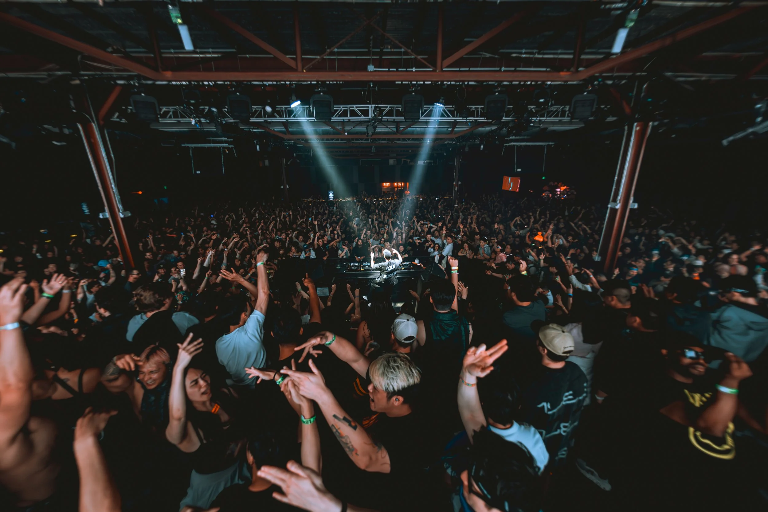 Wide-angle shot from behind the crowd of artist Audien performing at an indoor venue. A single bright spotlight illuminates the DJ booth as a massive crowd of fans raises their hands. Cinematic event photography with a dark, moody edit.
